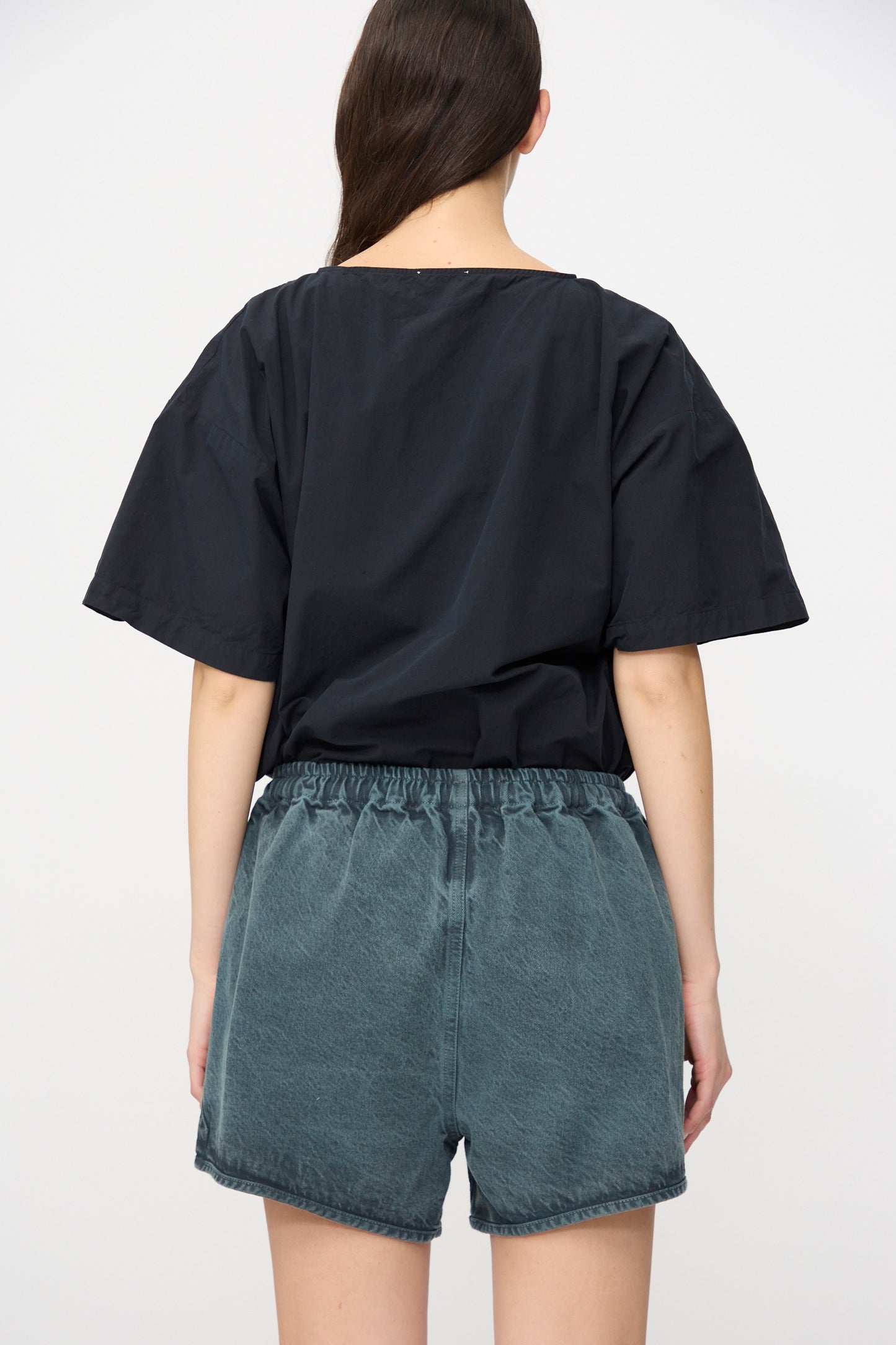 A person stands facing away from the camera, wearing a loose black short-sleeve top and SONO's high-waisted Schulz Shorts in Bluestone, UK-made denim, against a plain background.