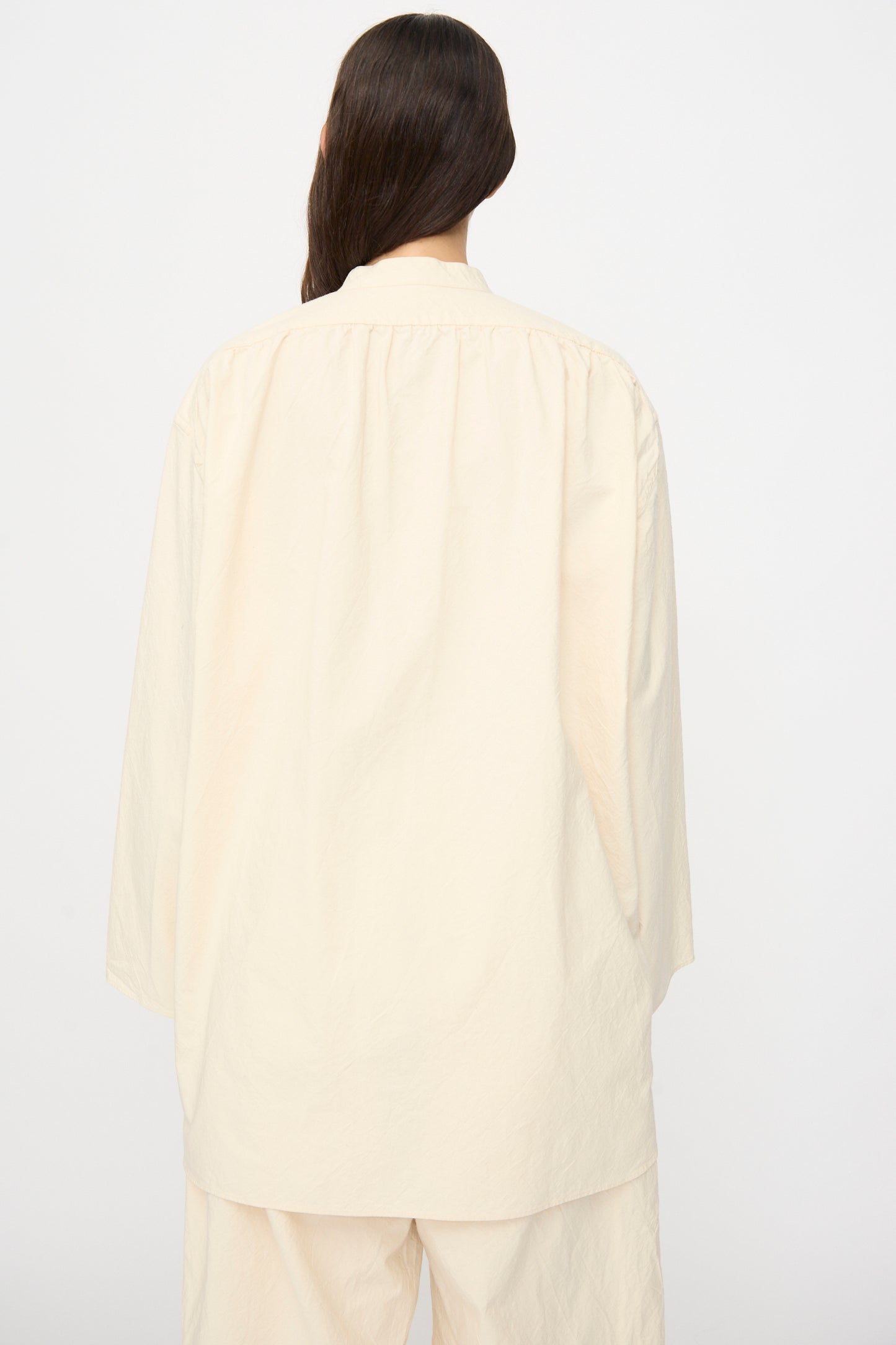 A person with long dark hair is shown from the back against a plain white background, wearing the SONO Shaman Shirt in Ecru—an oversized, long-sleeve organic cotton top paired with matching pants.