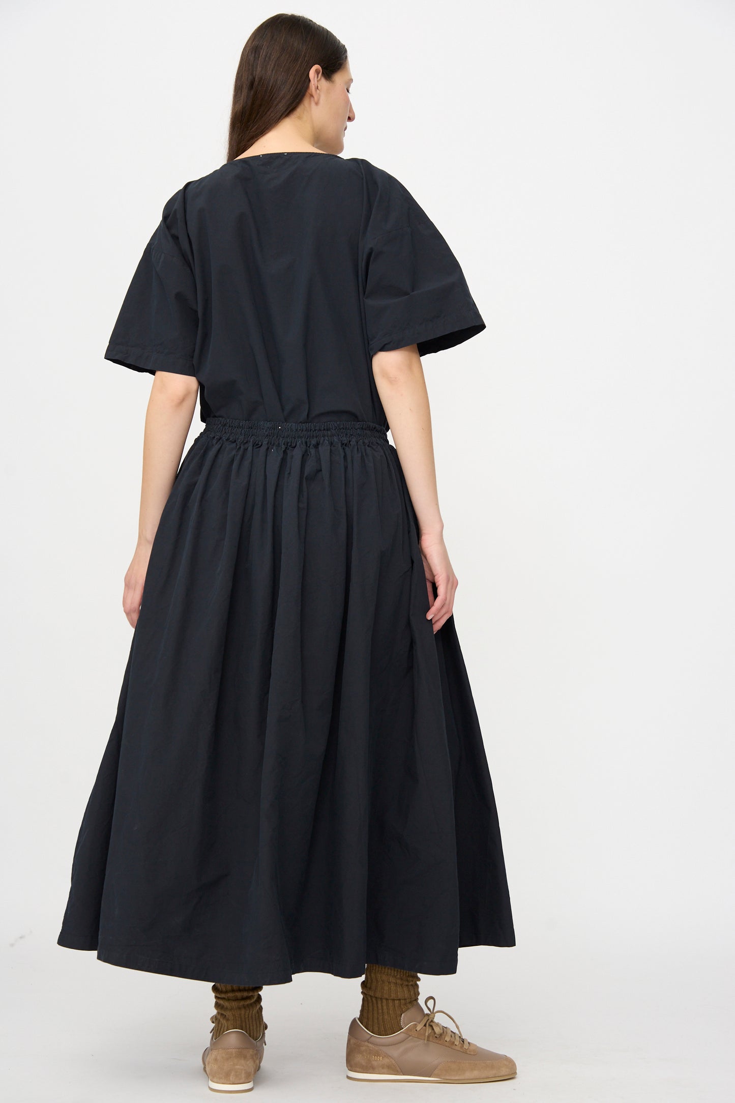 A person stands against a plain white background wearing the SONO Skye Skirt in Off Black—a loose, breezy linen cotton blend with a long A-line silhouette—paired with brown shoes and mid-calf socks.