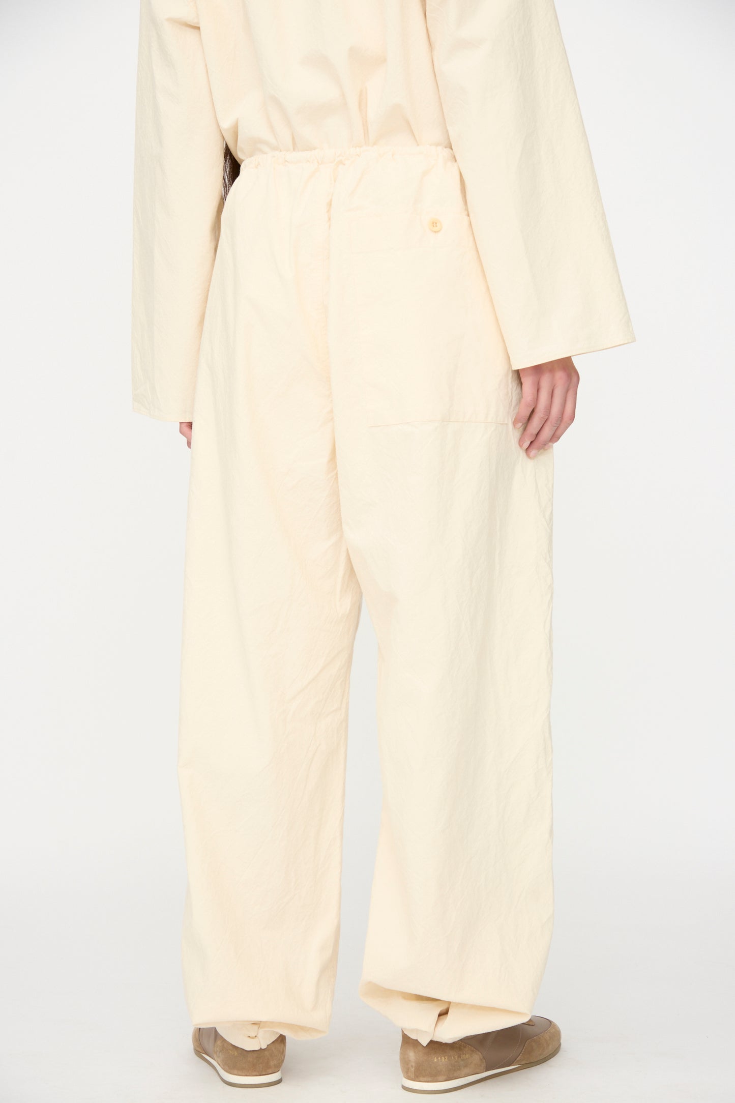 A person stands against a white background, shown from the back, wearing SONO's Sol Snowpant in Ecru—loose-fitting organic cotton pants with a drawstring waist and matching top—paired with tan shoes.