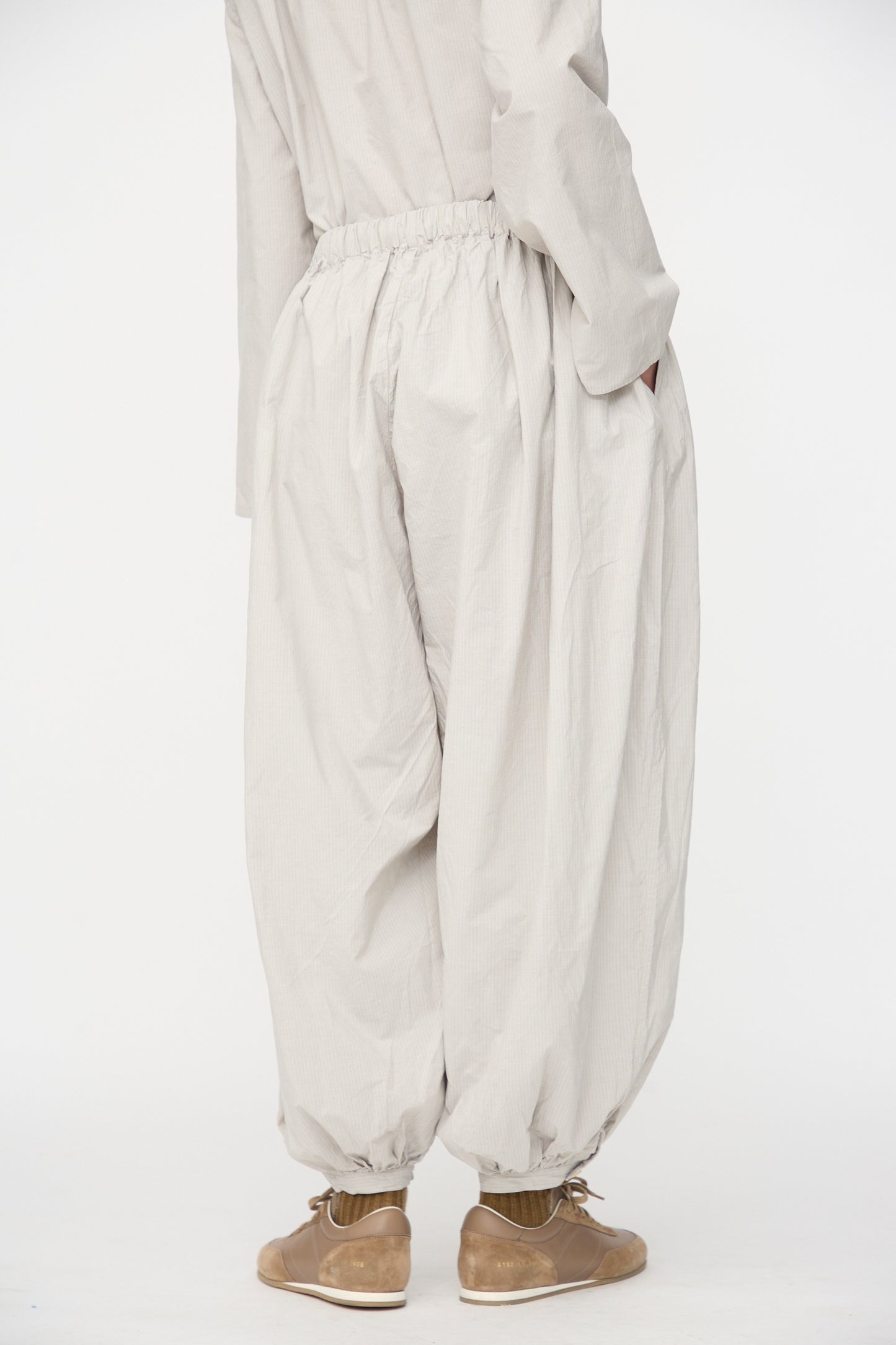 A person wears SONO’s Tamou Trousers in Dusty Stripe Fine—loose, light-colored pants with elastic cuffs—paired with tan shoes, standing against a plain white background.