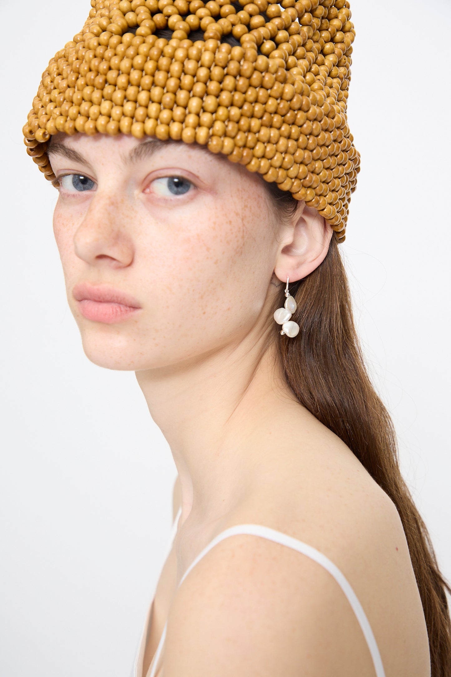 A fair-skinned, freckled woman poses against a plain white background wearing a tan beaded hat, white strappy top, and the Santangelo Baba Single Earring in Triple Baby.