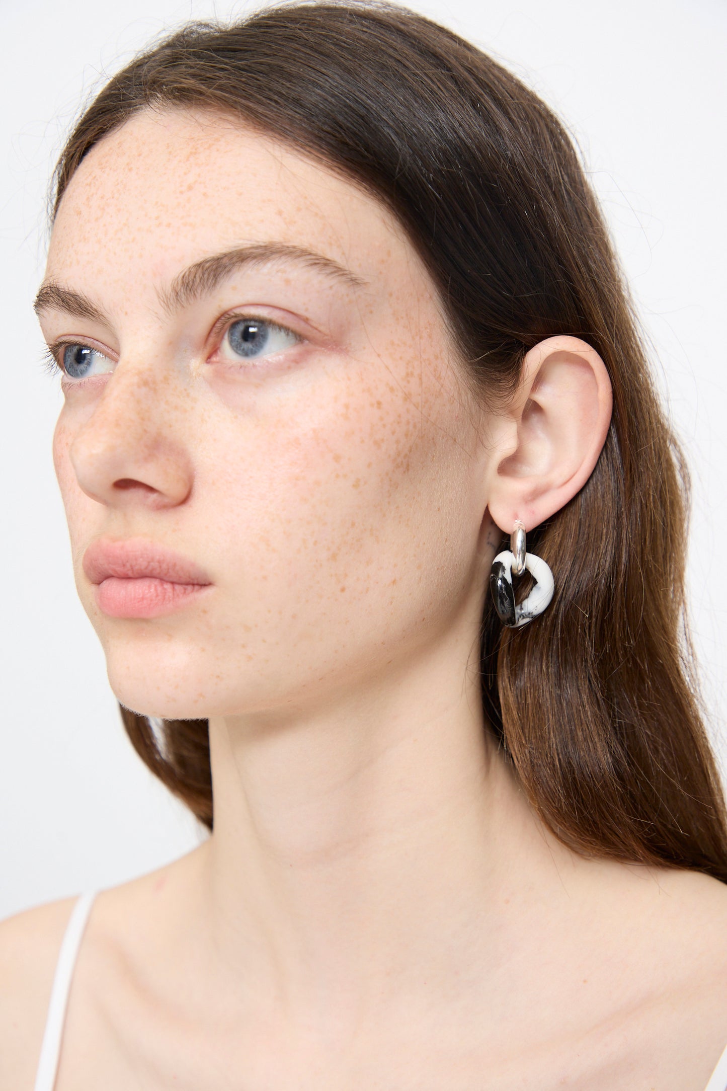 A woman with long brown hair wears Santangelo’s Bad Orb Earrings in Zebra Jasper—chunky, linked black-and-white hoops. Shown from the shoulders up against a plain background, these striking earrings are handmade in NYC.