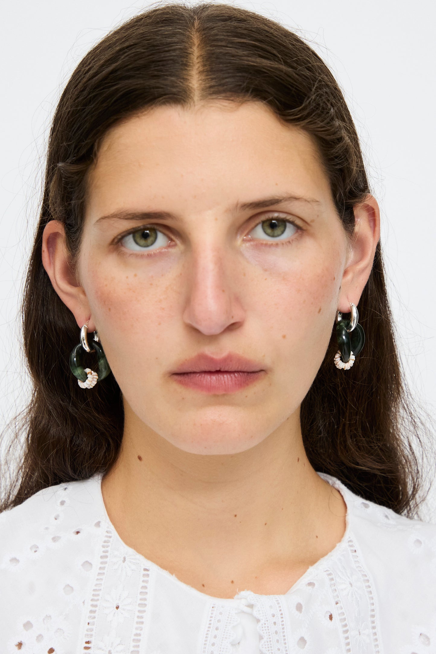 A woman with long brown hair, wearing a white eyelet blouse and Santangelo Bad Orb Earrings in Jade, looks directly at the camera with a neutral expression.