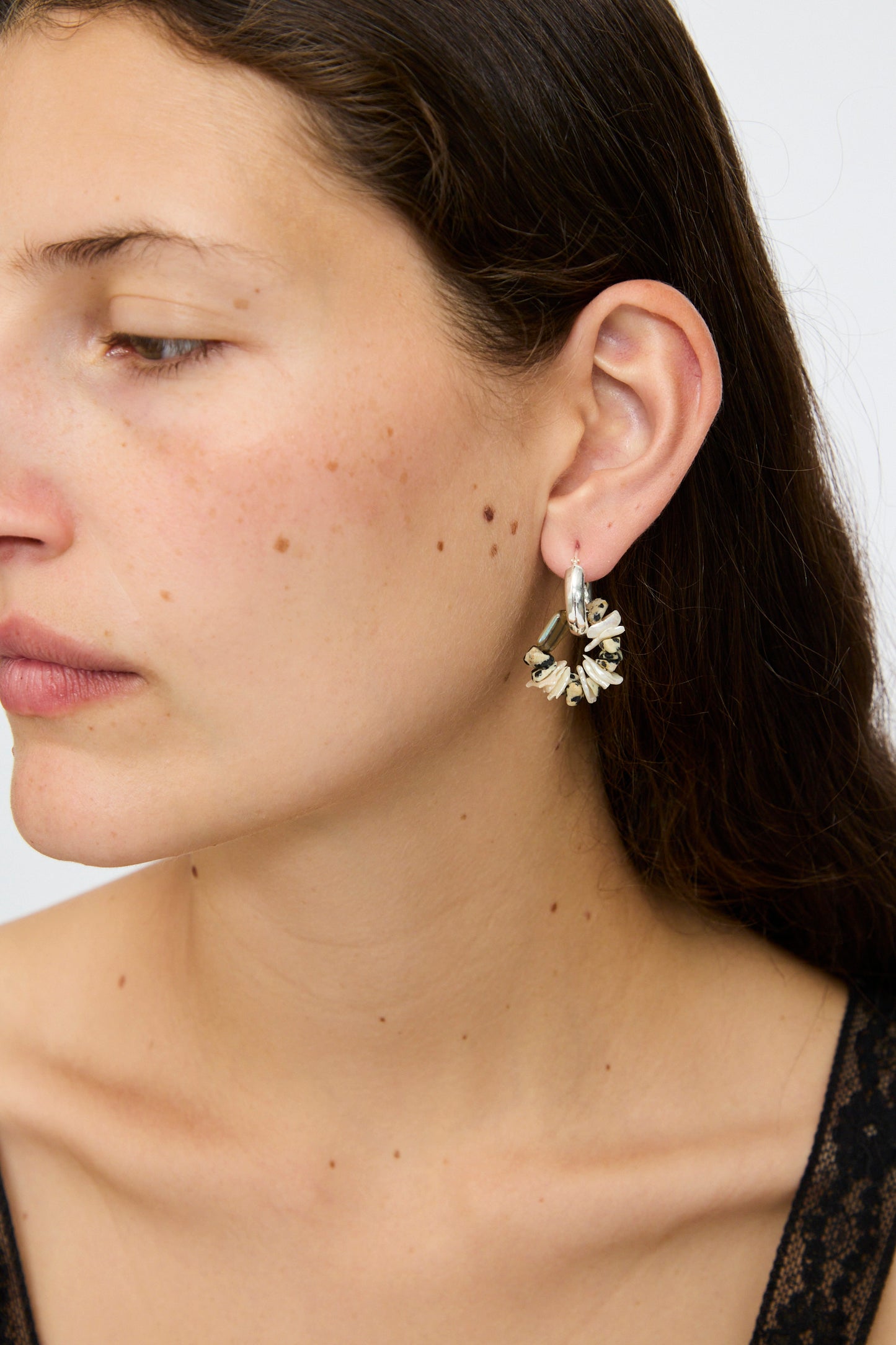 A person with long brown hair wears Santangelo's Che Bomba Earrings in Abalone Hoop, featuring silver hoops adorned with small white flowers and freshwater cornflake pearls, shown from the side against a plain background.