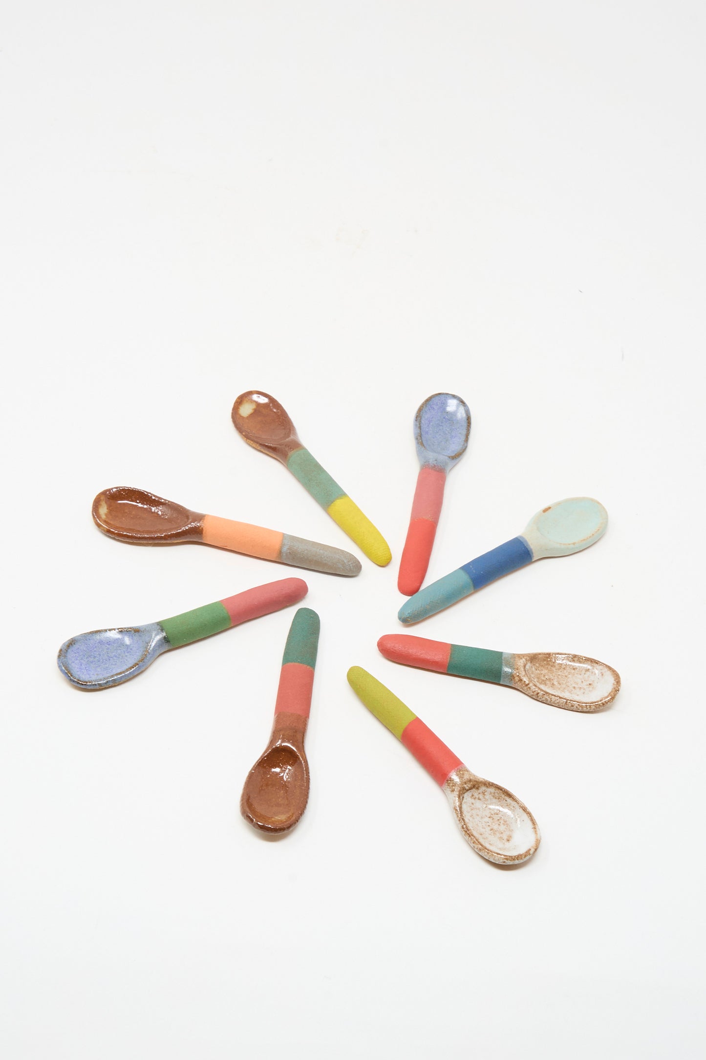 Nine Shino Takeda Condiment Spoons in Grey Orange are arranged in a circle on a white background.