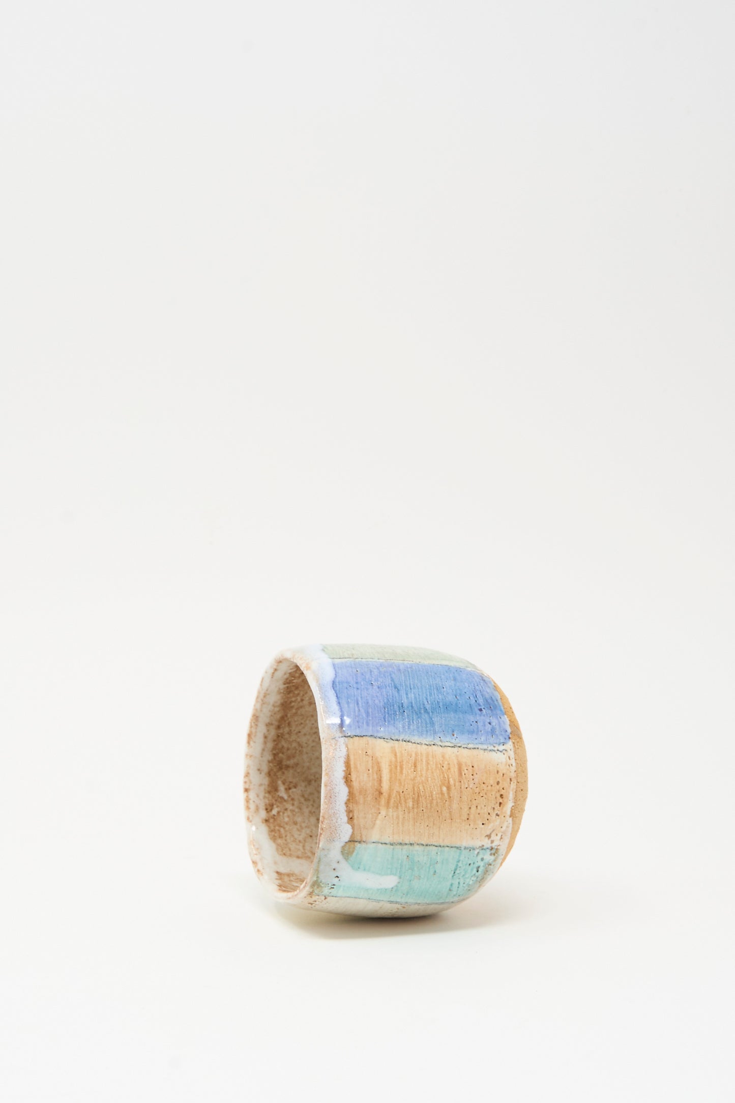 The Cup in Stripe by Shino Takeda is a small cylindrical ceramic cup featuring hand-painted blue, tan, and turquoise vertical stripes on a textured surface, shown against a plain white background.