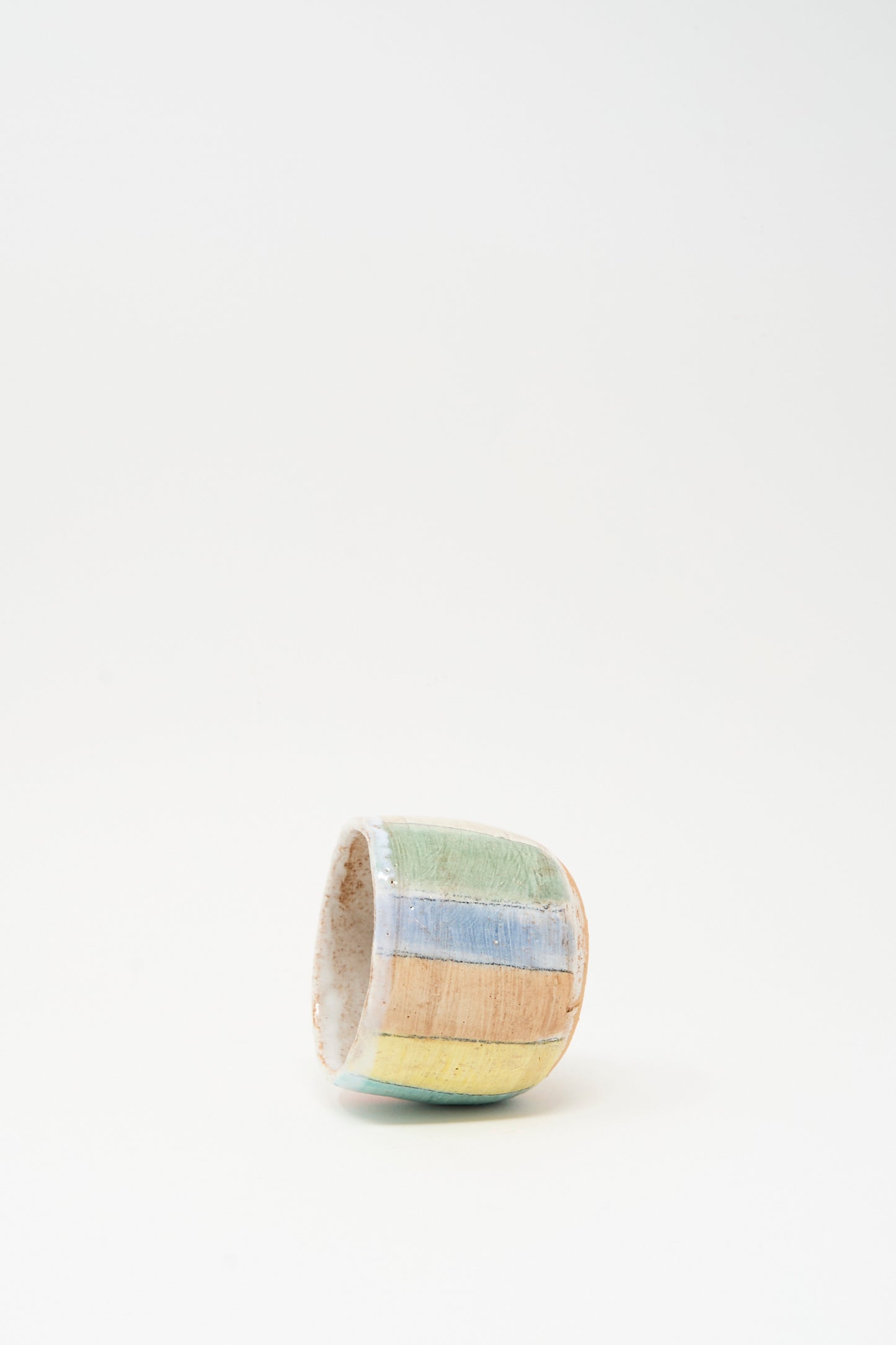 Cup in Stripe I by Shino Takeda is a small hand-painted ceramic cup with horizontal pastel green, blue, yellow, and orange stripes, made in New York and shown on its side against a plain white background.