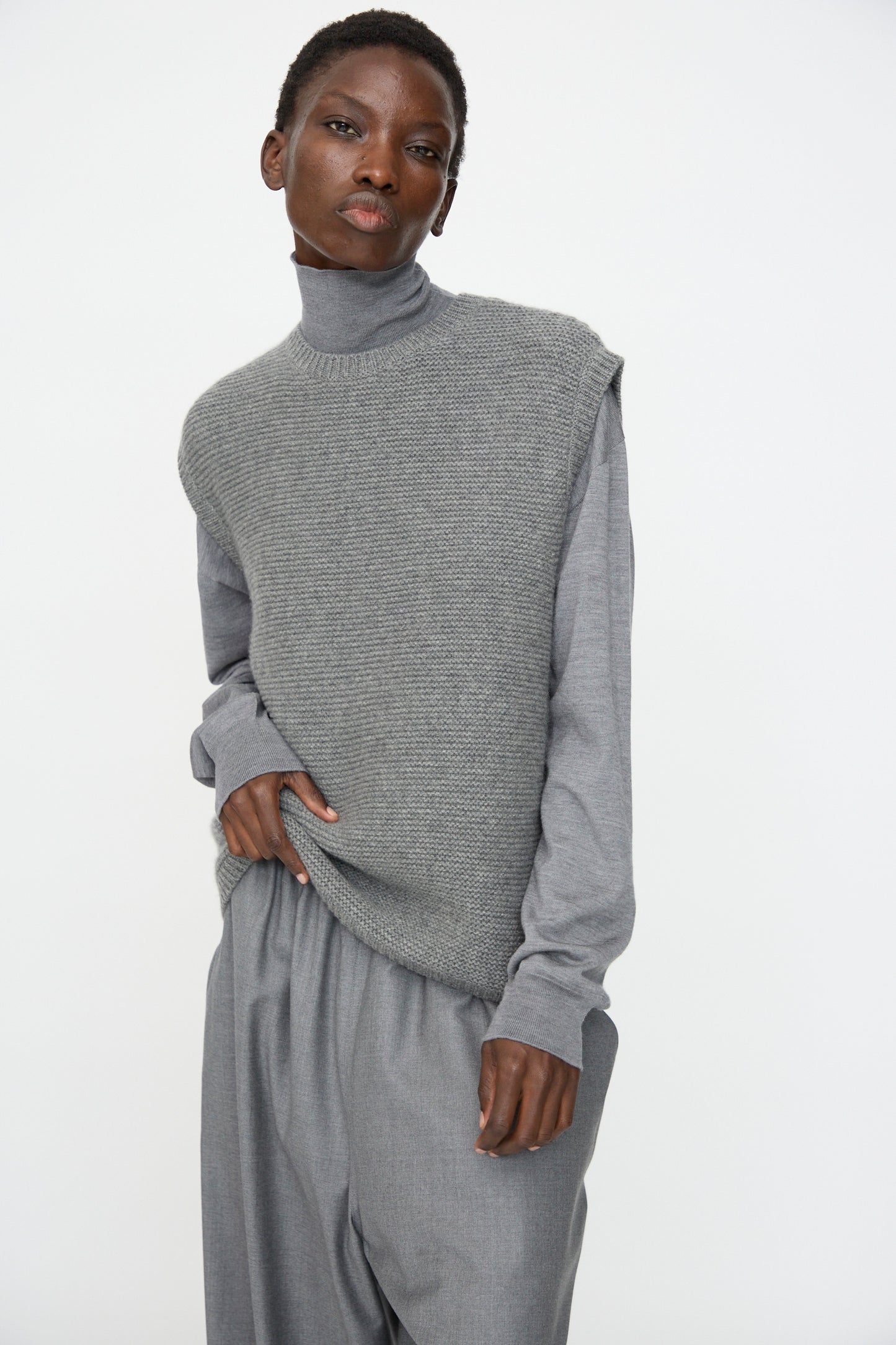 A person wears the Sofie D'Hoore Cashmere Matta Sweater in Mid Grey Melange, a sleeveless textured vest, and loose grey pants, standing against a plain white background.