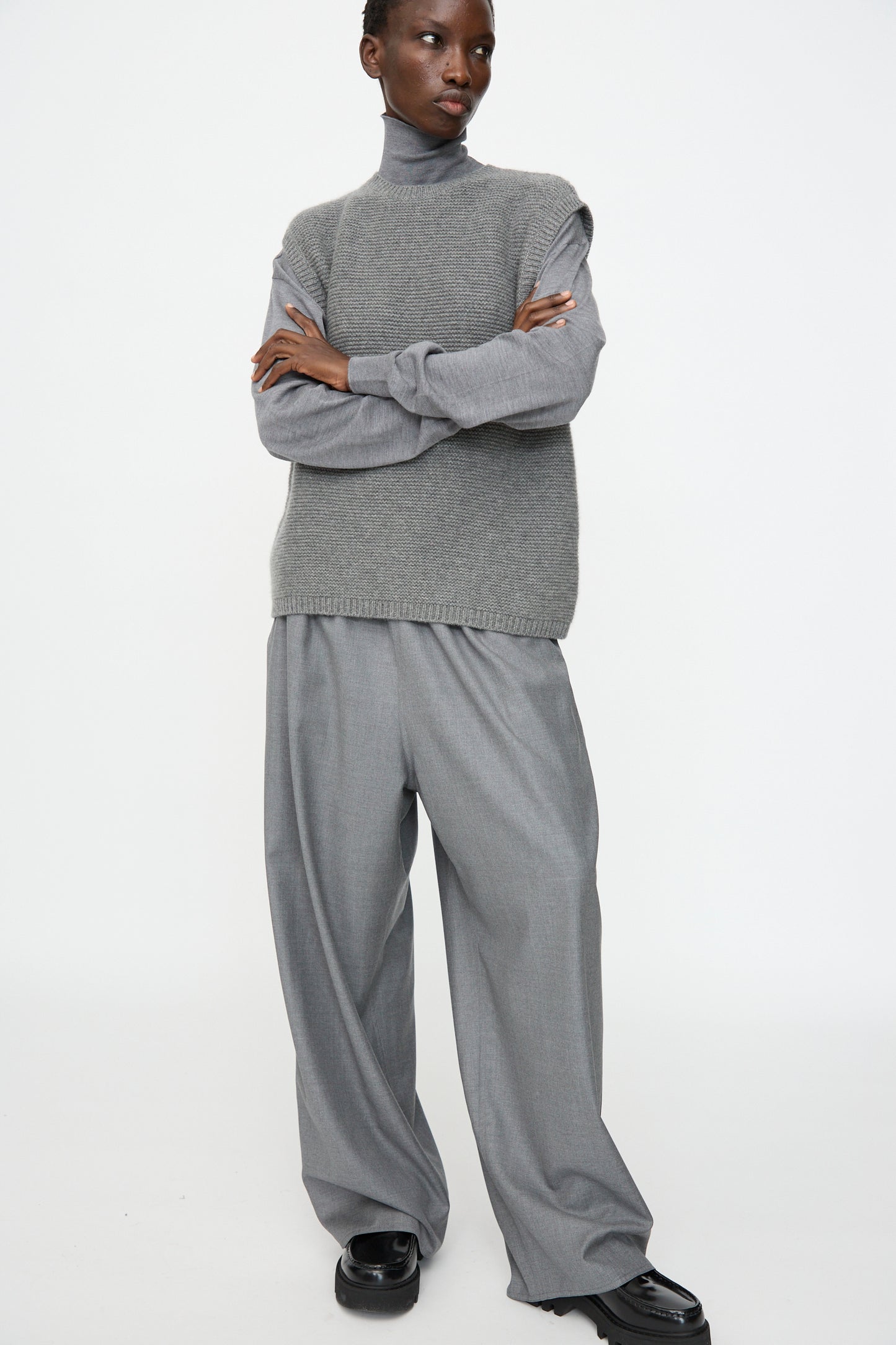 Person stands with arms crossed, wearing the Sofie D'Hoore Cashmere Matta Sweater in Mid Grey Melange over a gray turtleneck, paired with wide-leg trousers and black shoes against a plain white background.