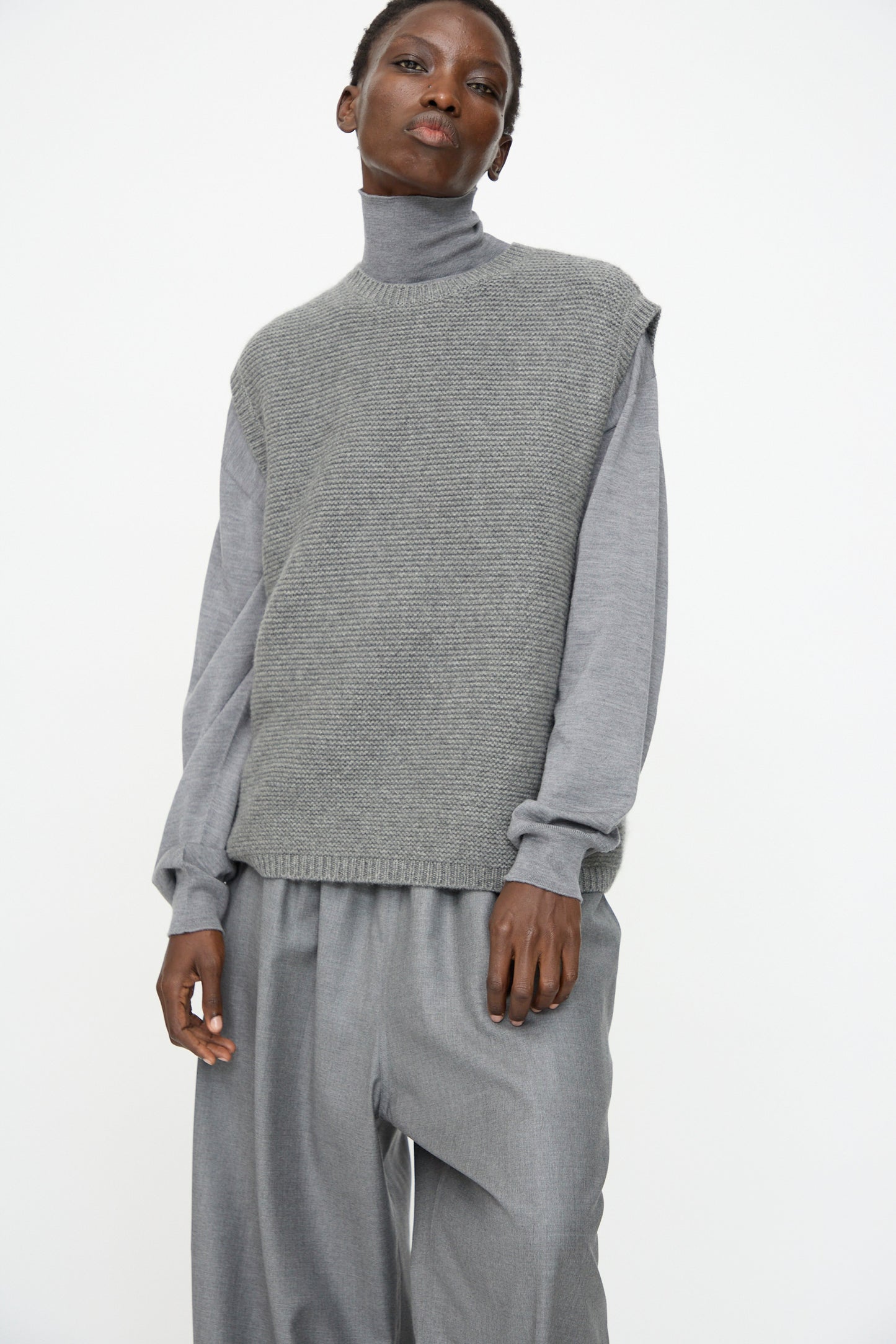 A person wears Sofie D'Hoore's Cashmere Matta Sweater in Mid Grey Melange over a grey turtleneck and loose grey trousers, standing against a plain white background.