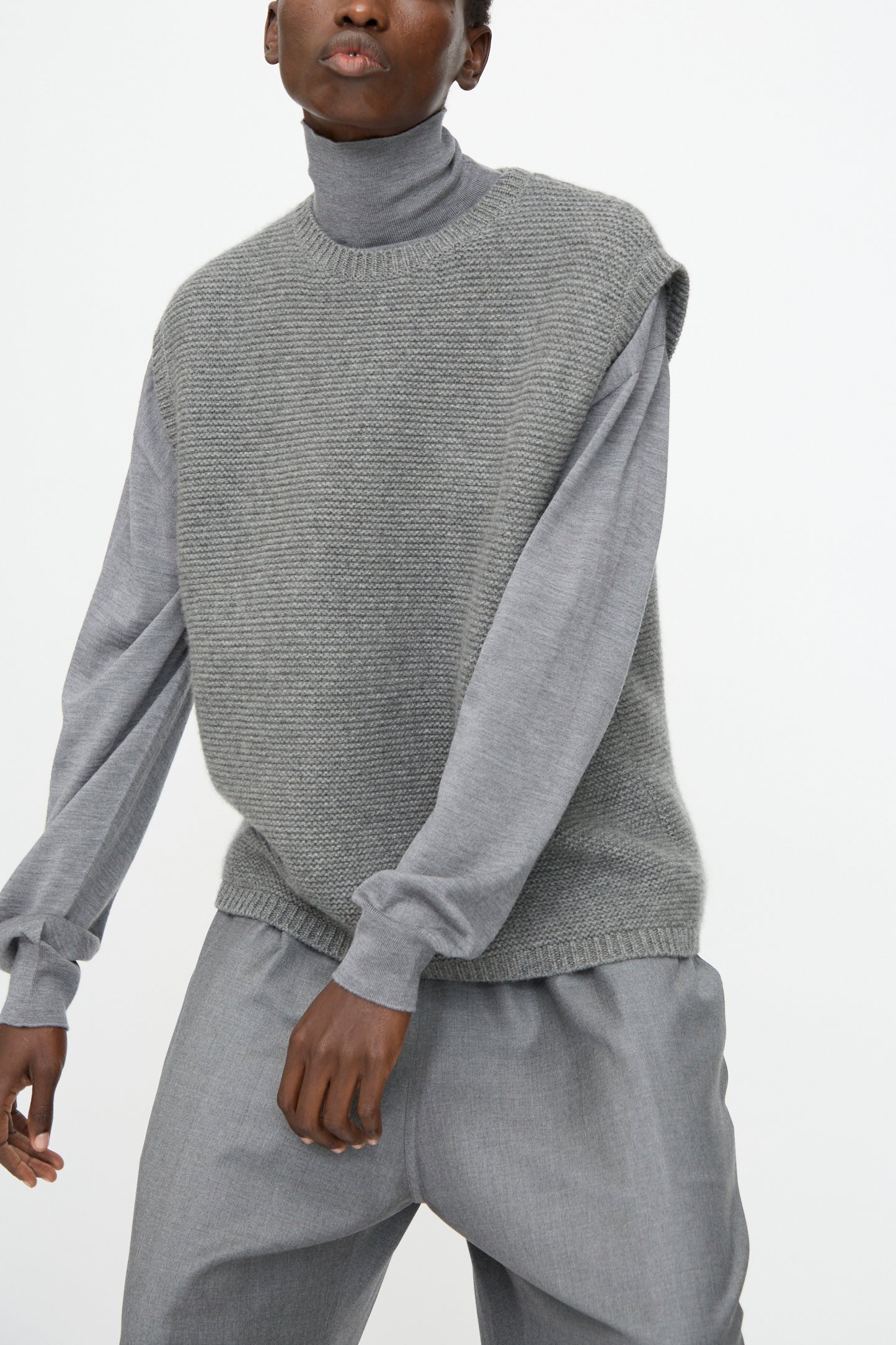 Model wearing the Sofie D'Hoore Cashmere Matta Sweater in Mid Grey Melange under a sleeveless knit vest with loose grey pants, posed against a plain white background.