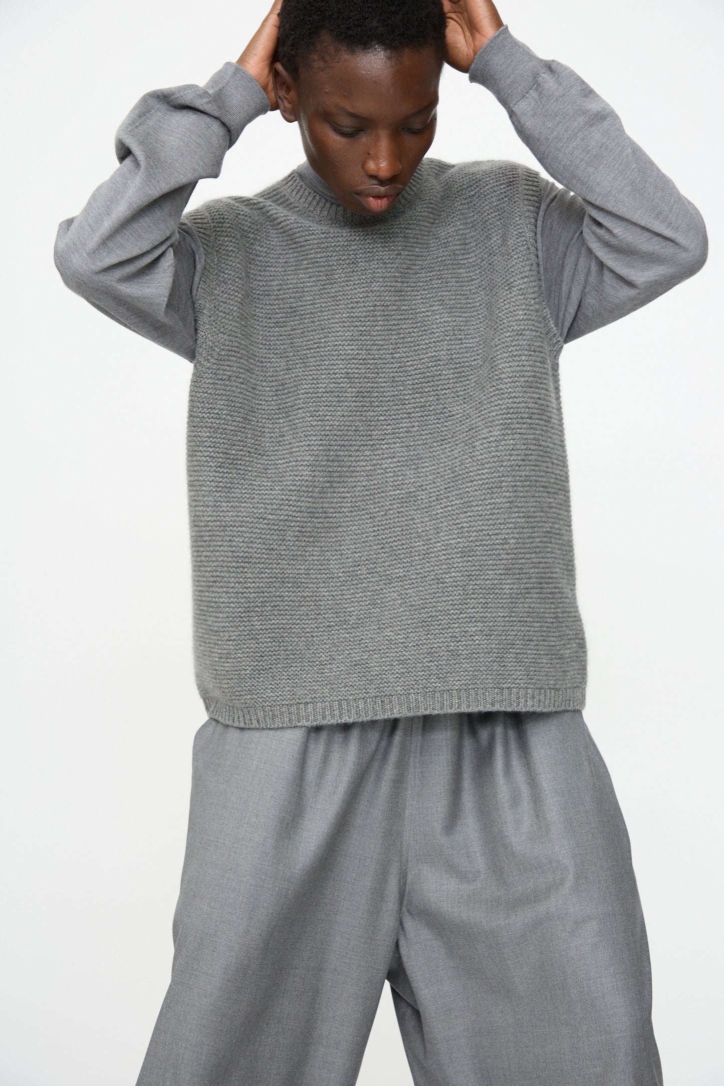 A person wears the Sofie D'Hoore Cashmere Matta Sweater in Mid Grey Melange over a long-sleeved shirt and wide-leg gray pants, standing against a plain light background with hands on their head.