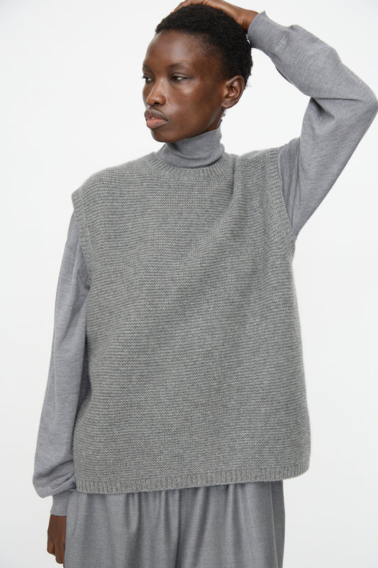 A person wearing the Sofie D'Hoore Cashmere Matta Sweater in Mid Grey Melange, a textured grey sleeveless vest, and gray pants stands with one arm raised against a plain white background.