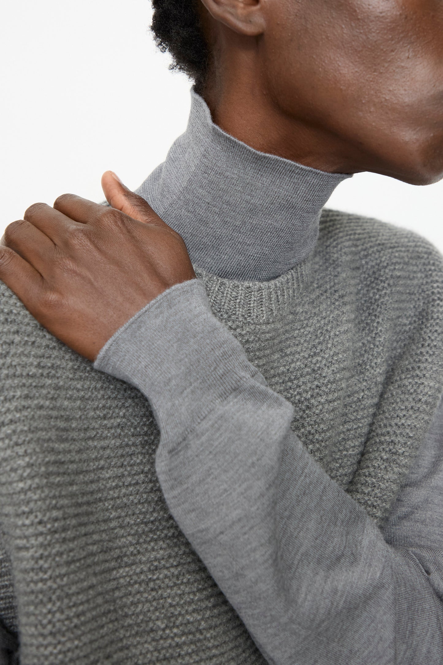 A person wears the Sofie D'Hoore Cashmere Matta Sweater in Mid Grey Melange over a light gray turtleneck, with one hand resting on their shoulder.