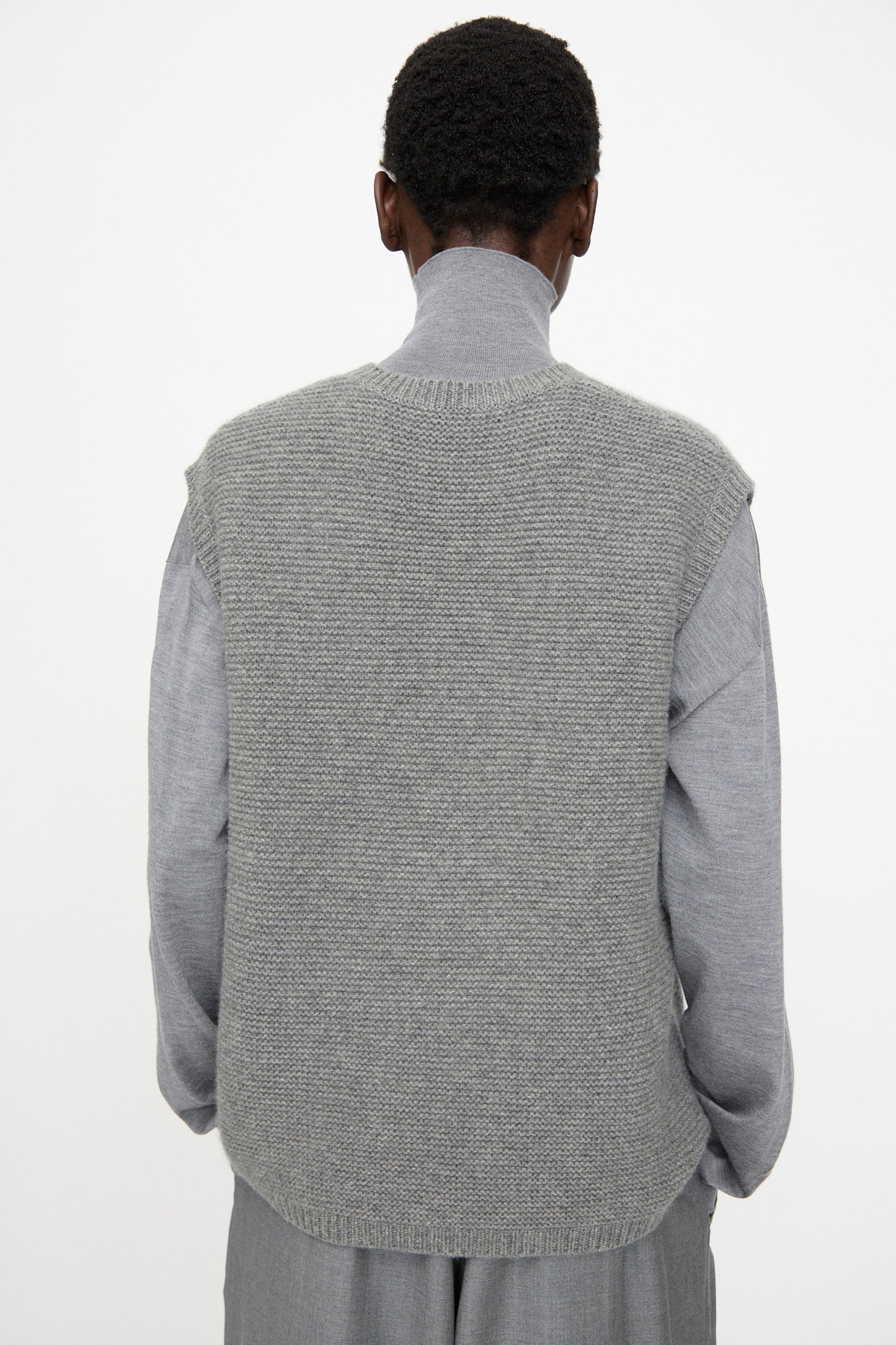 A person stands with their back to the camera, wearing the Sofie D'Hoore Cashmere Matta Sweater in Mid Grey Melange over a light gray long-sleeve shirt and gray pants, set against a plain light background.