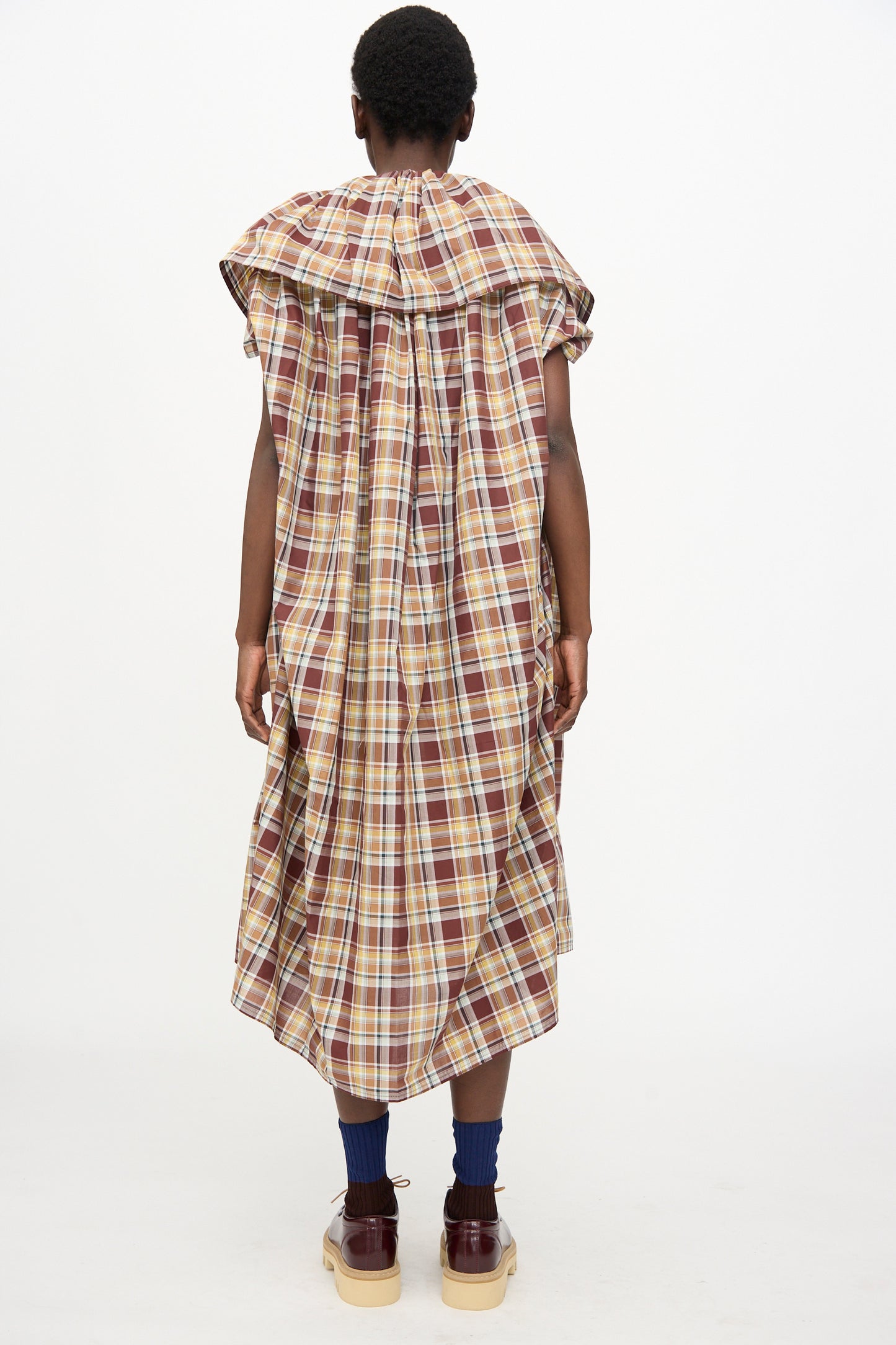 A person stands facing away, wearing the Sofie D'Hoore Cotton Batiste Check Dolabella Dress in Toffee, paired with blue socks and brown platform shoes.