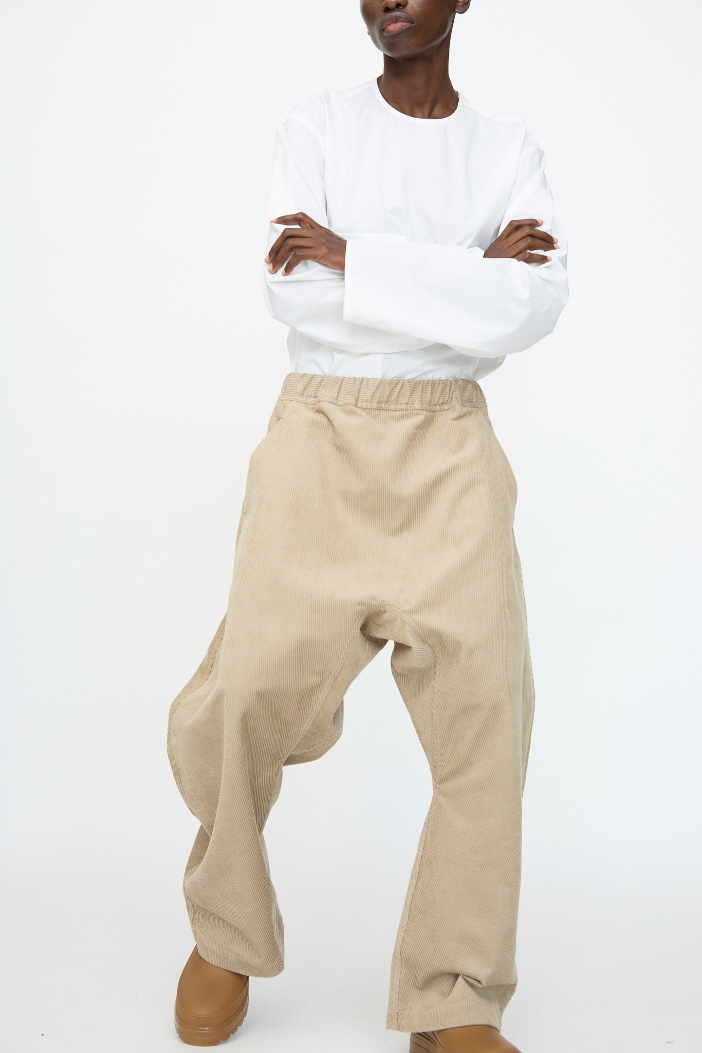 A person stands with arms crossed against a white background, wearing a loose white long-sleeve shirt and Sofie D’Hoore’s Cotton Corduroy Plof Pant in Cashew matched with tan shoes.
