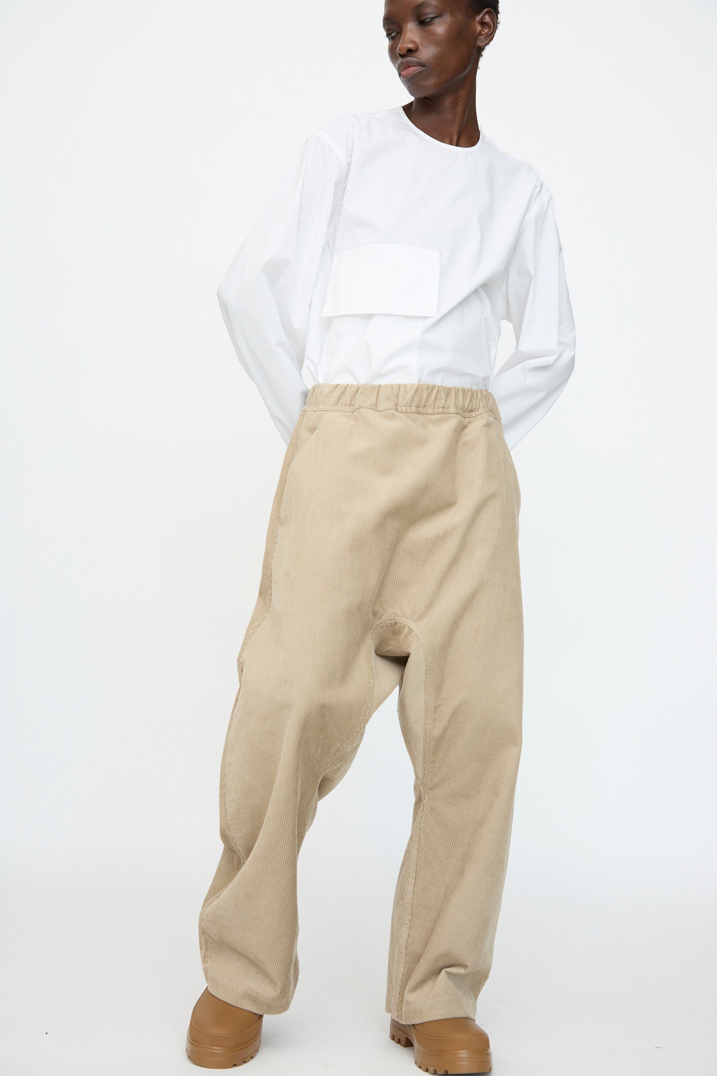A person stands against a plain white background wearing a white long-sleeve top, Sofie D'Hoore Cotton Corduroy Plof Pant in Cashew, and tan platform shoes.