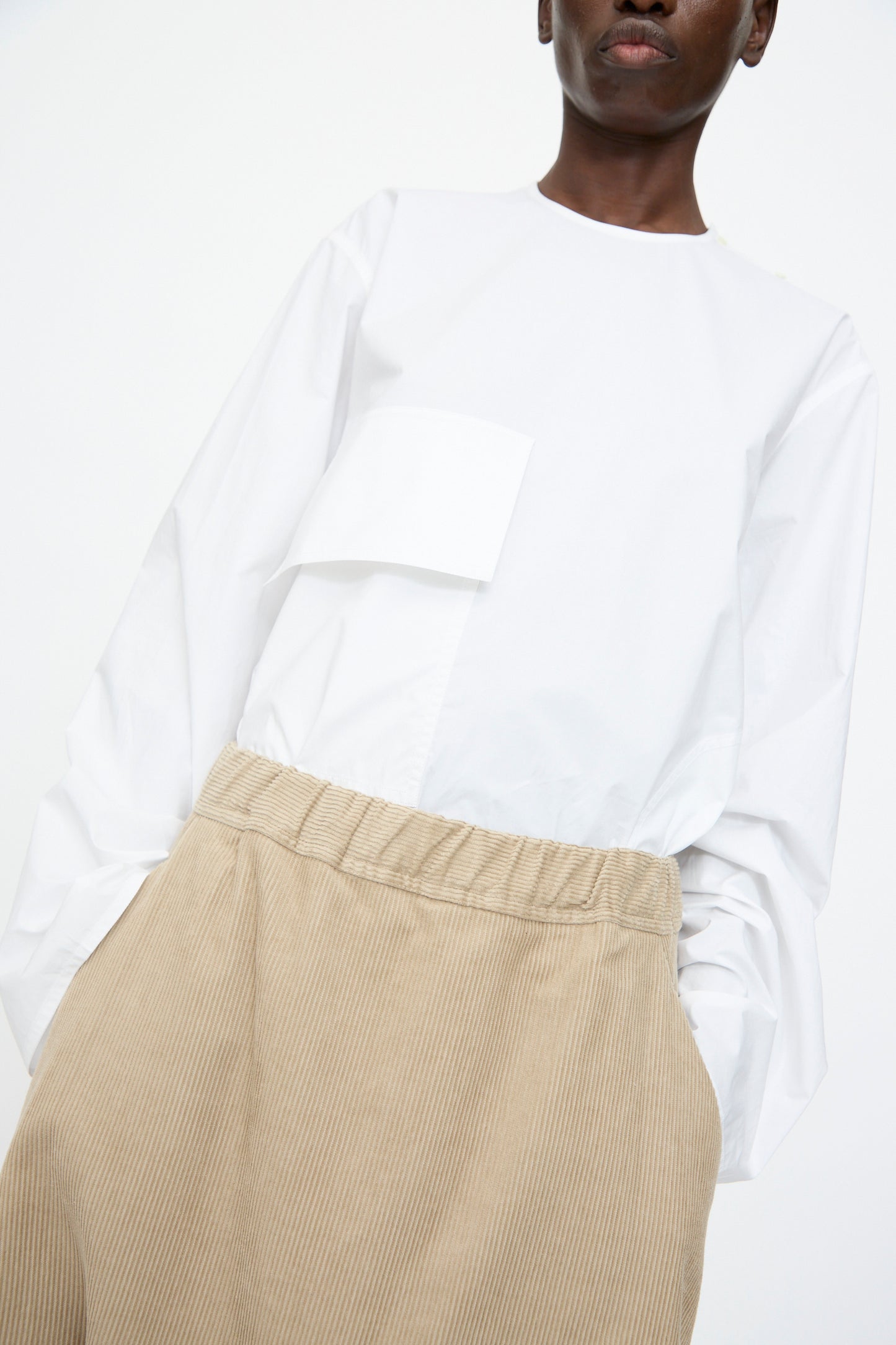 A person wears Sofie D'Hoore's Cotton Corduroy Plof Pant in Cashew—relaxed-fit beige corduroy pants with an elastic waistband—paired with a loose white long-sleeve top featuring a square pocket, standing against a plain background.