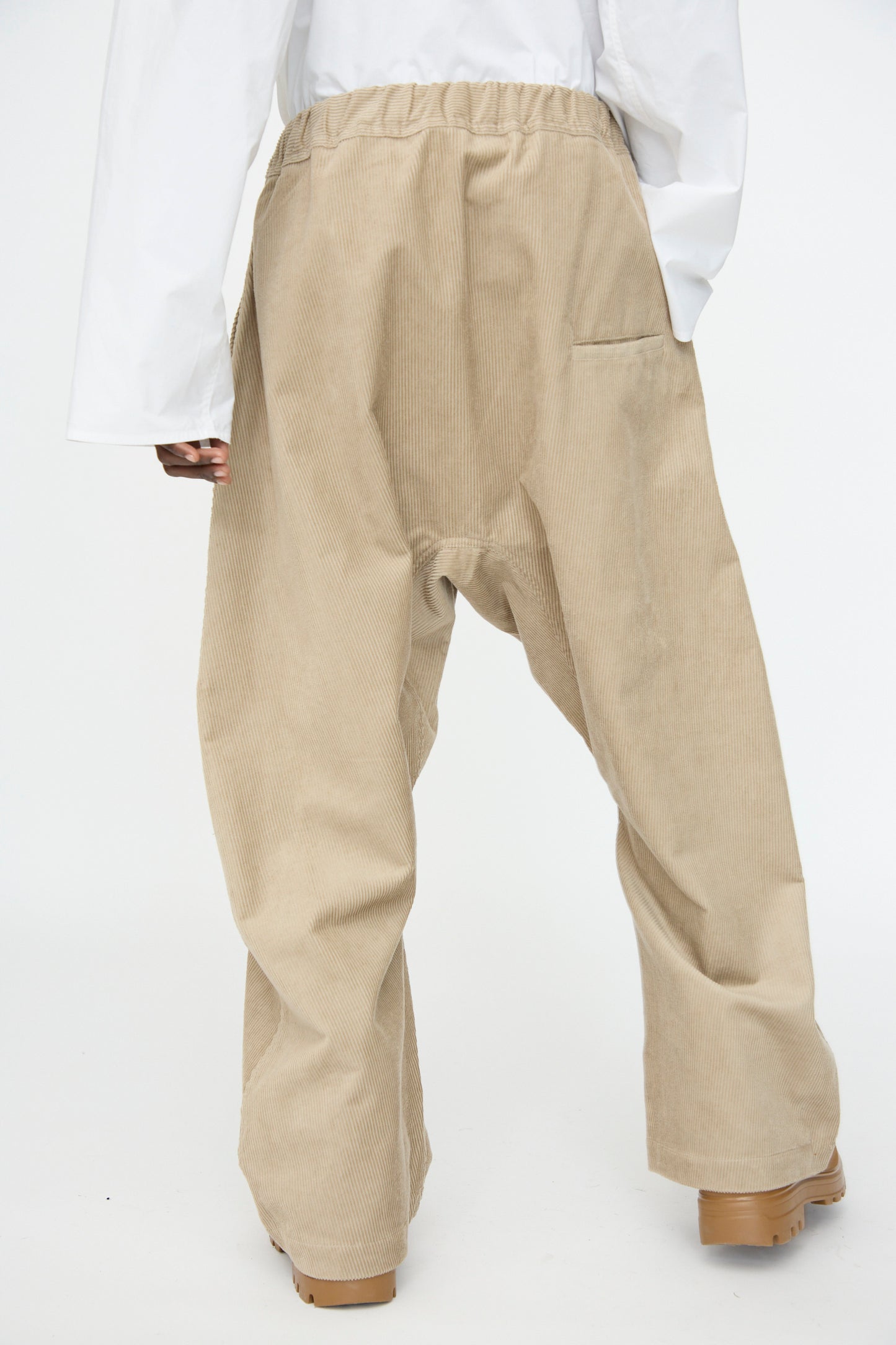 Model wears Sofie D'Hoore's Cotton Corduroy Plof Pant in Cashew, featuring a relaxed fit and elastic waistband, paired with a white long-sleeve shirt. Photographed from the back against a plain background.