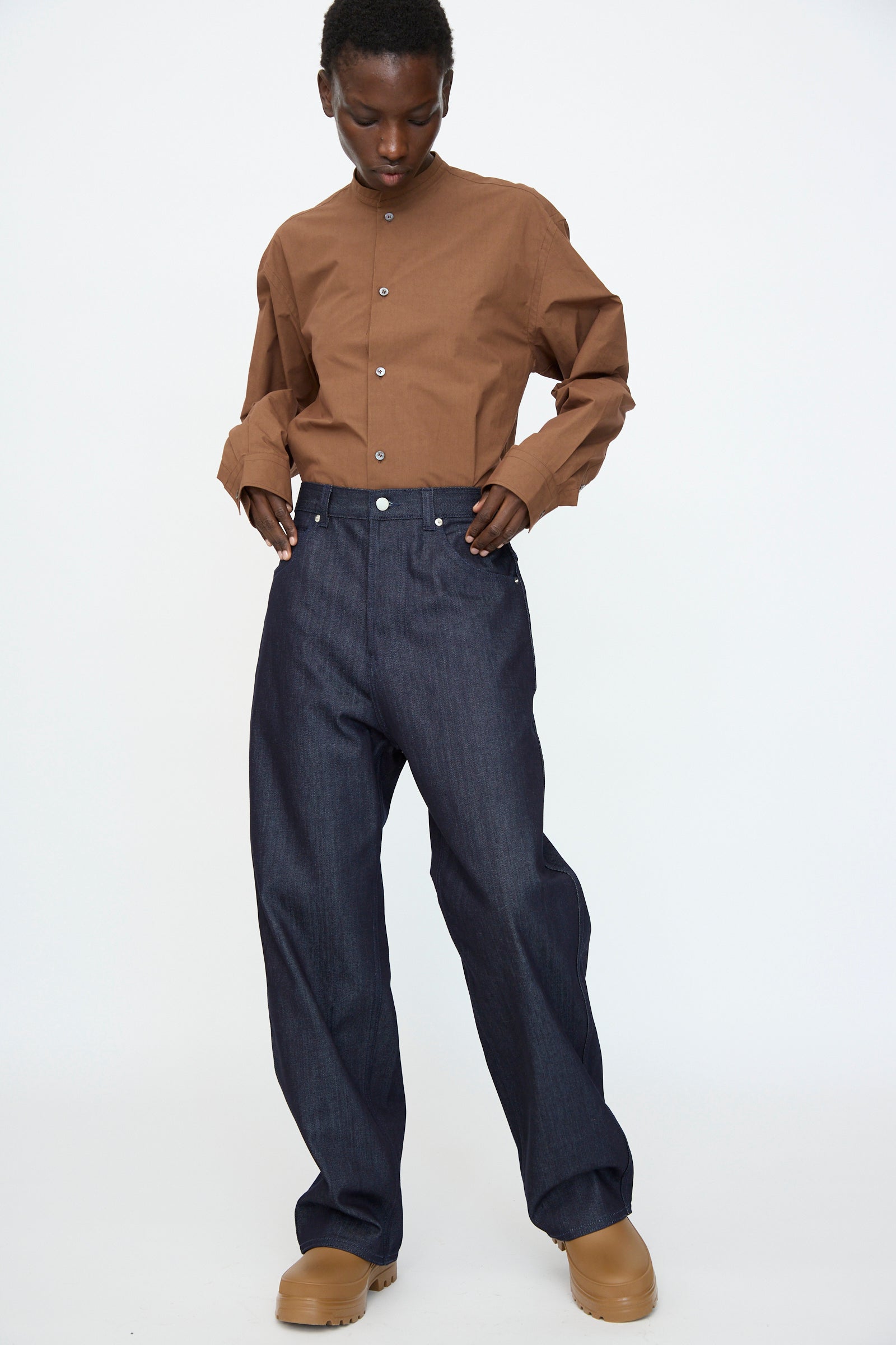 Person standing against a plain background, wearing the Sofie D’Hoore Denim Peggy Pant in Raw—loose, wide-leg 5-pocket jeans—and a brown button-up shirt, with tan shoes, looking down with hands tucked in pockets.