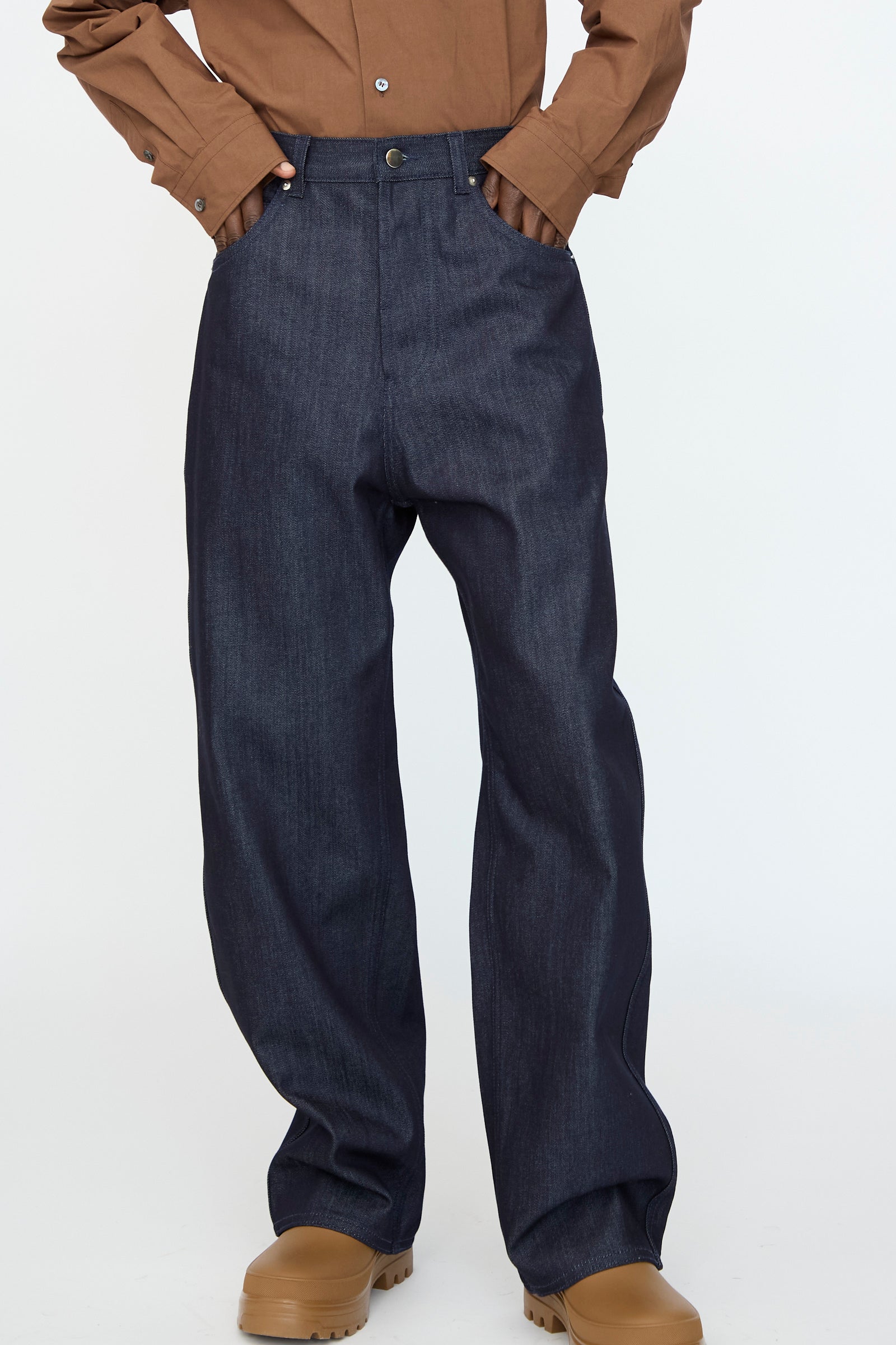 A person wearing the Sofie D’Hoore Denim Peggy Pant in Raw, styled with a brown button-up shirt and tan boots, stands against a plain white background.