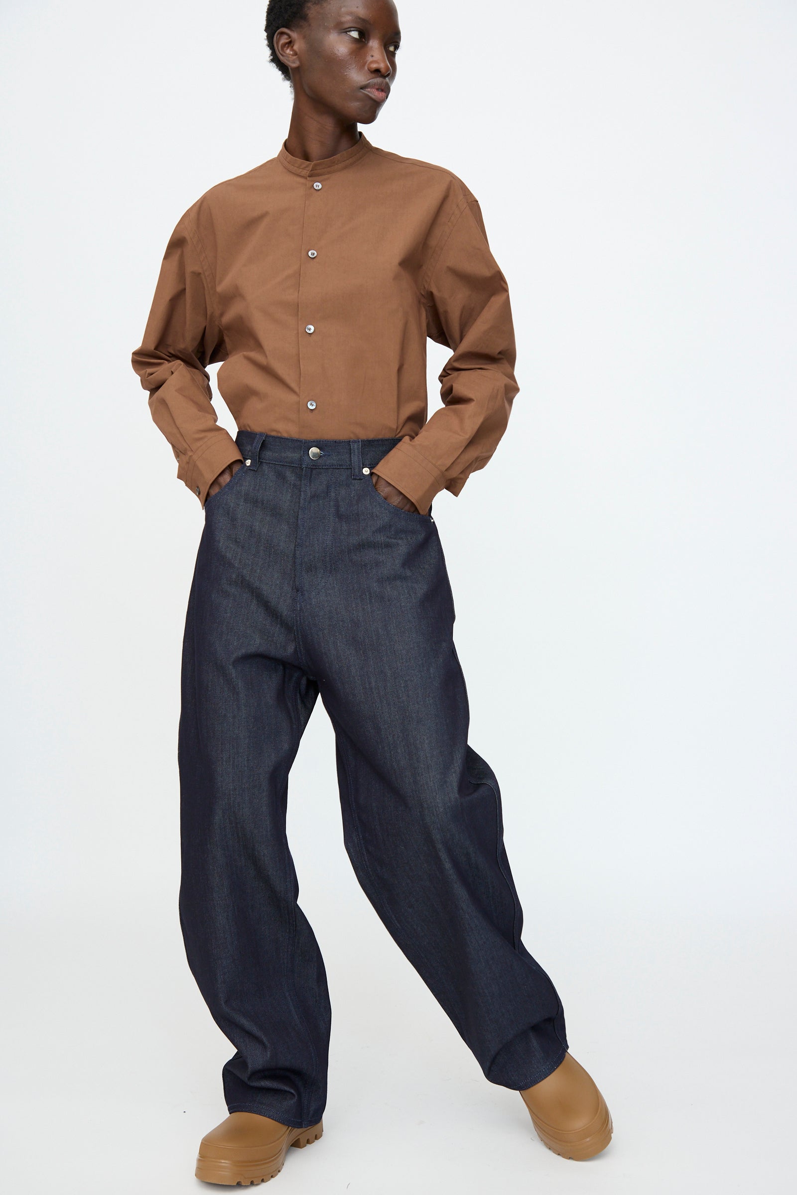A person wearing a brown button-up shirt, Sofie D'Hoore Denim Peggy Pant in Raw, and tan shoes stands against a plain white background.