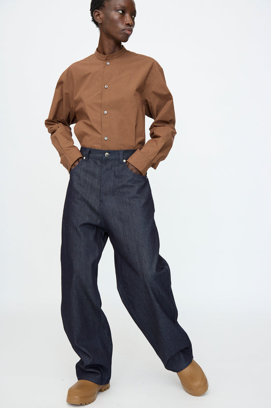 A person wearing a brown button-up shirt, Sofie D'Hoore Denim Peggy Pant in Raw, and tan shoes stands against a plain white background.