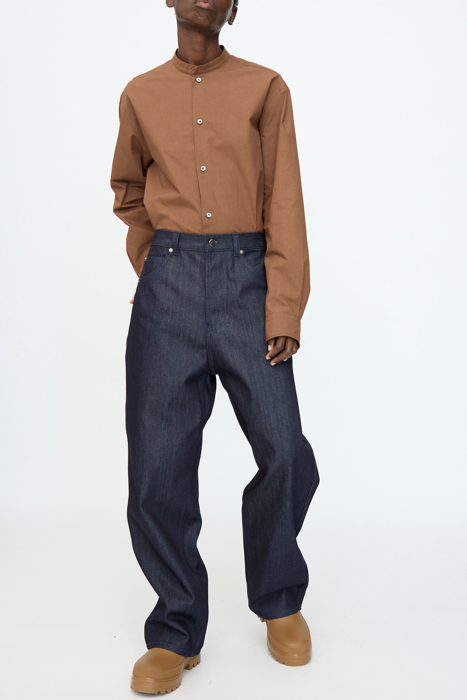 Person stands against a white background wearing a brown button-up shirt, Sofie D'Hoore Denim Peggy Pant in Raw, and tan shoes.