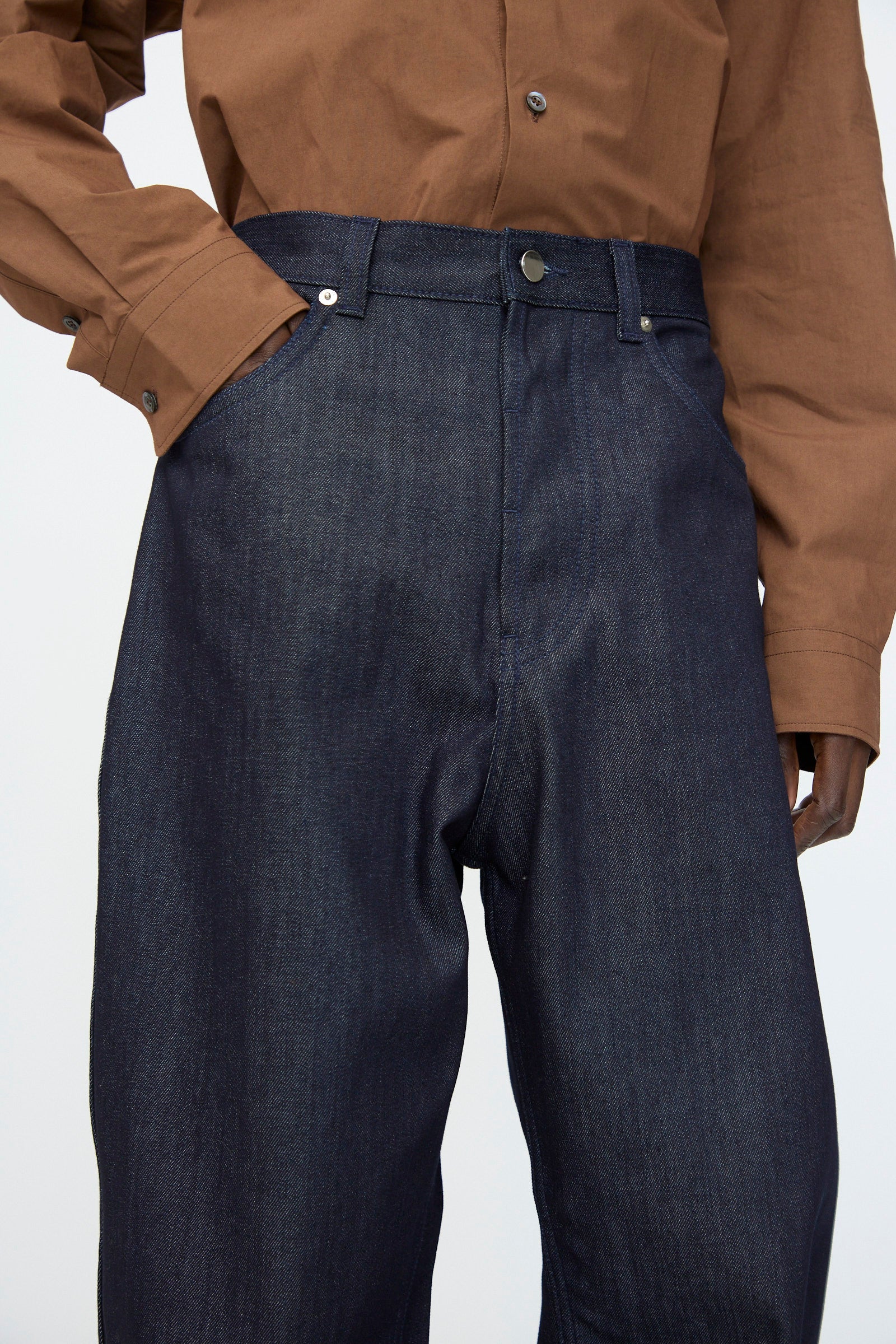 A person wears the Denim Peggy Pant in Raw by Sofie D'Hoore, a high-waisted, dark blue 5-pocket denim, paired with a brown button-up shirt and one hand in the pocket.