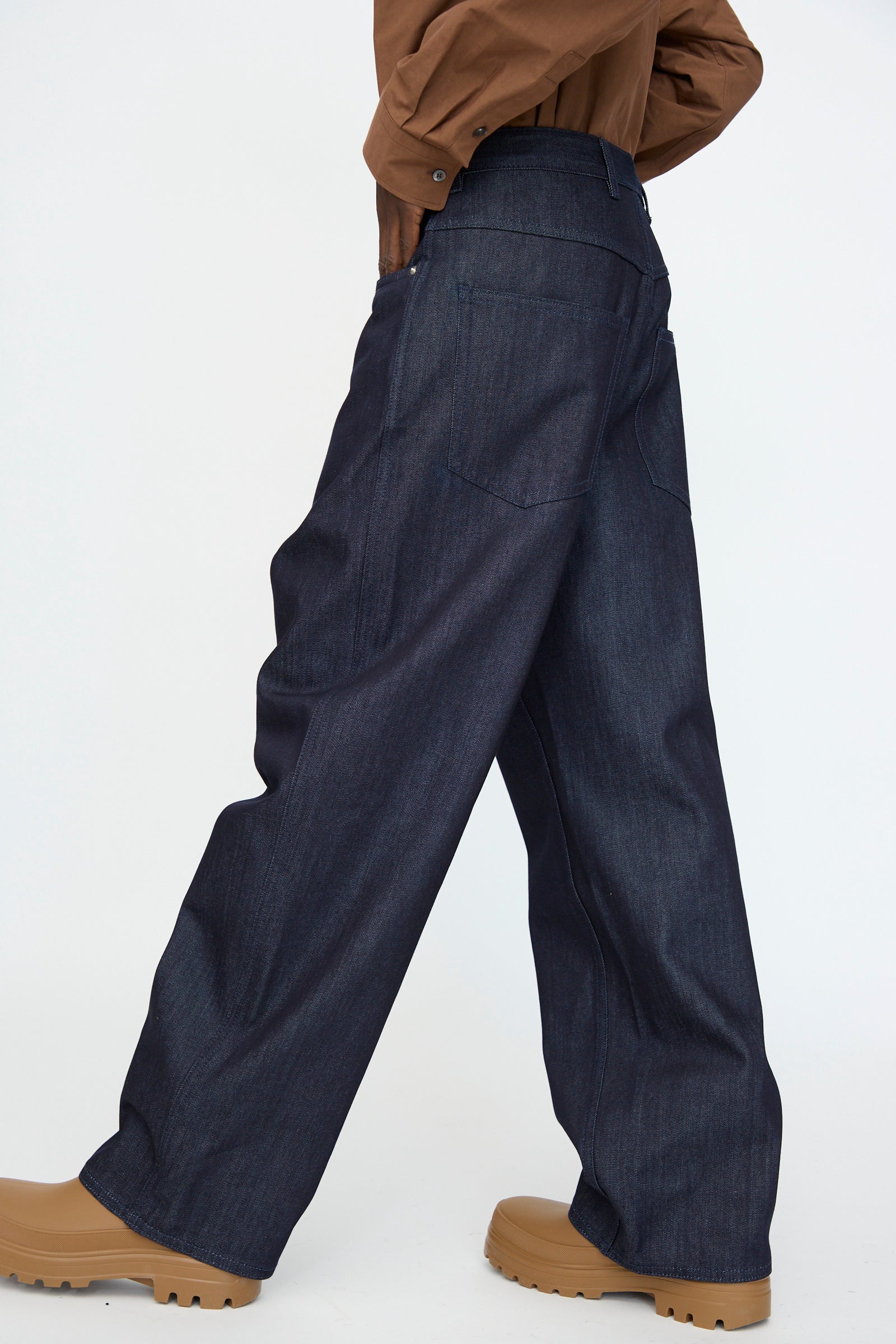 Model wears the Sofie D'Hoore Denim Peggy Pant in Raw—wide-leg dark denim with a classic 5-pocket design—paired with a brown long-sleeve shirt and tan chunky-soled shoes, photographed from the side against a plain background.