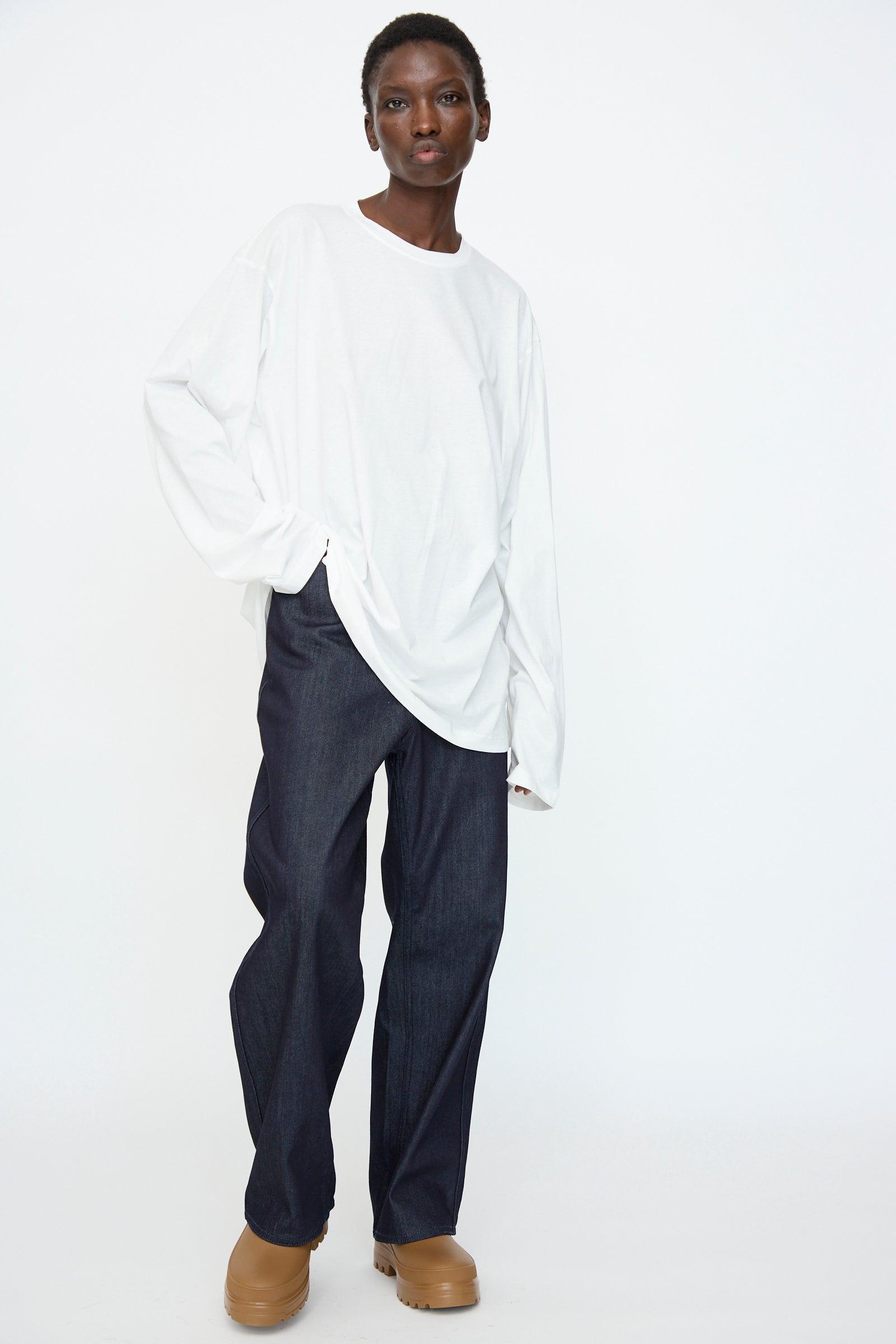 A person wearing the Sofie D'Hoore Fine Cotton Jersey Toledo Tee in Off White stands against a plain white background, paired with dark wide-leg jeans and tan shoes.