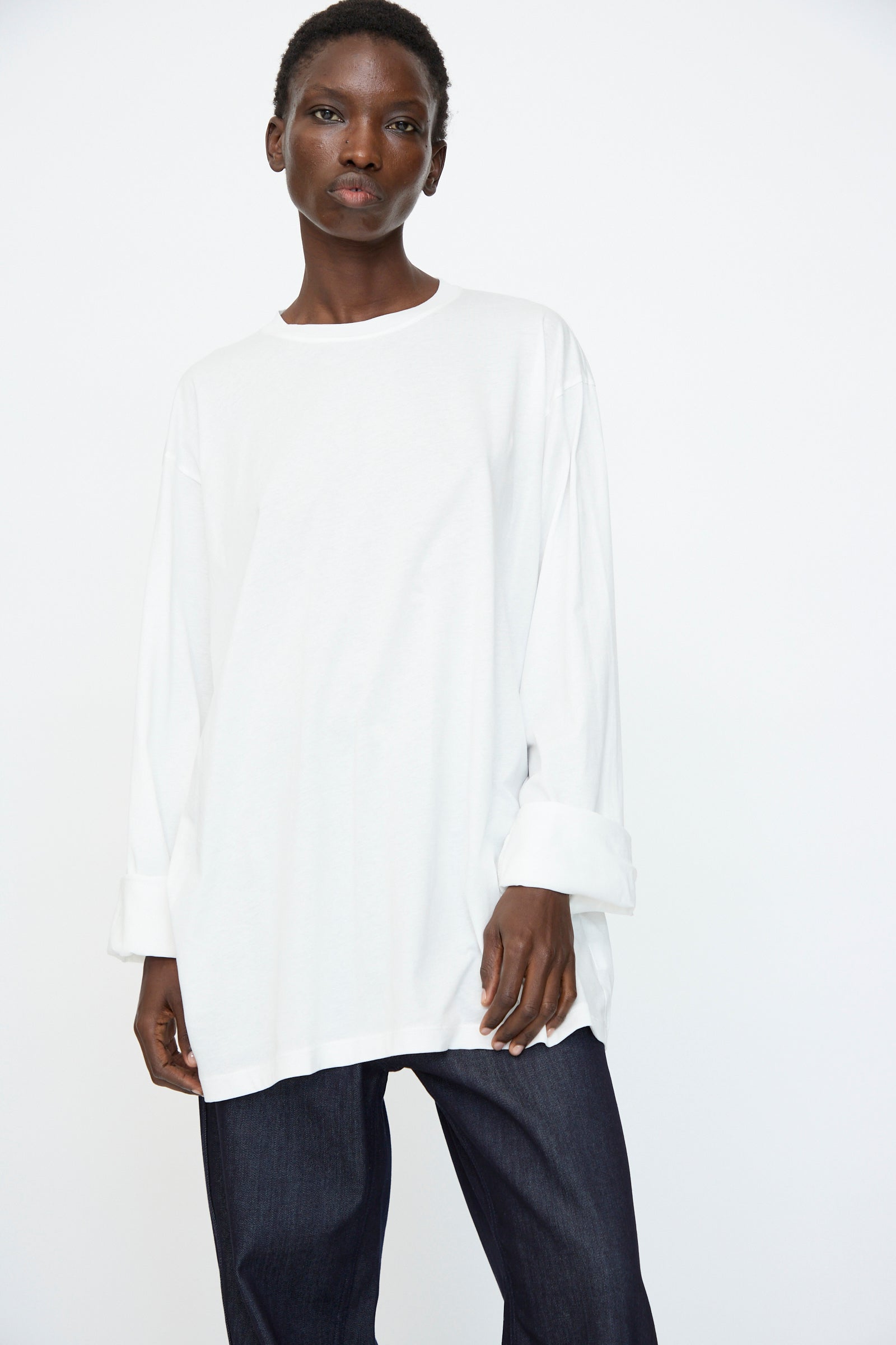 A person stands against a plain background wearing the Sofie D'Hoore Fine Cotton Jersey Toledo Tee in Off White and dark jeans, gazing directly at the camera.