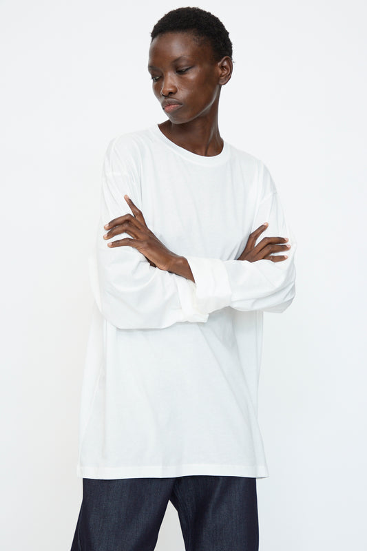 A person with short hair, arms crossed, wears the Sofie D'Hoore Fine Cotton Jersey Toledo Tee in Off White and dark pants, standing against a plain white background.