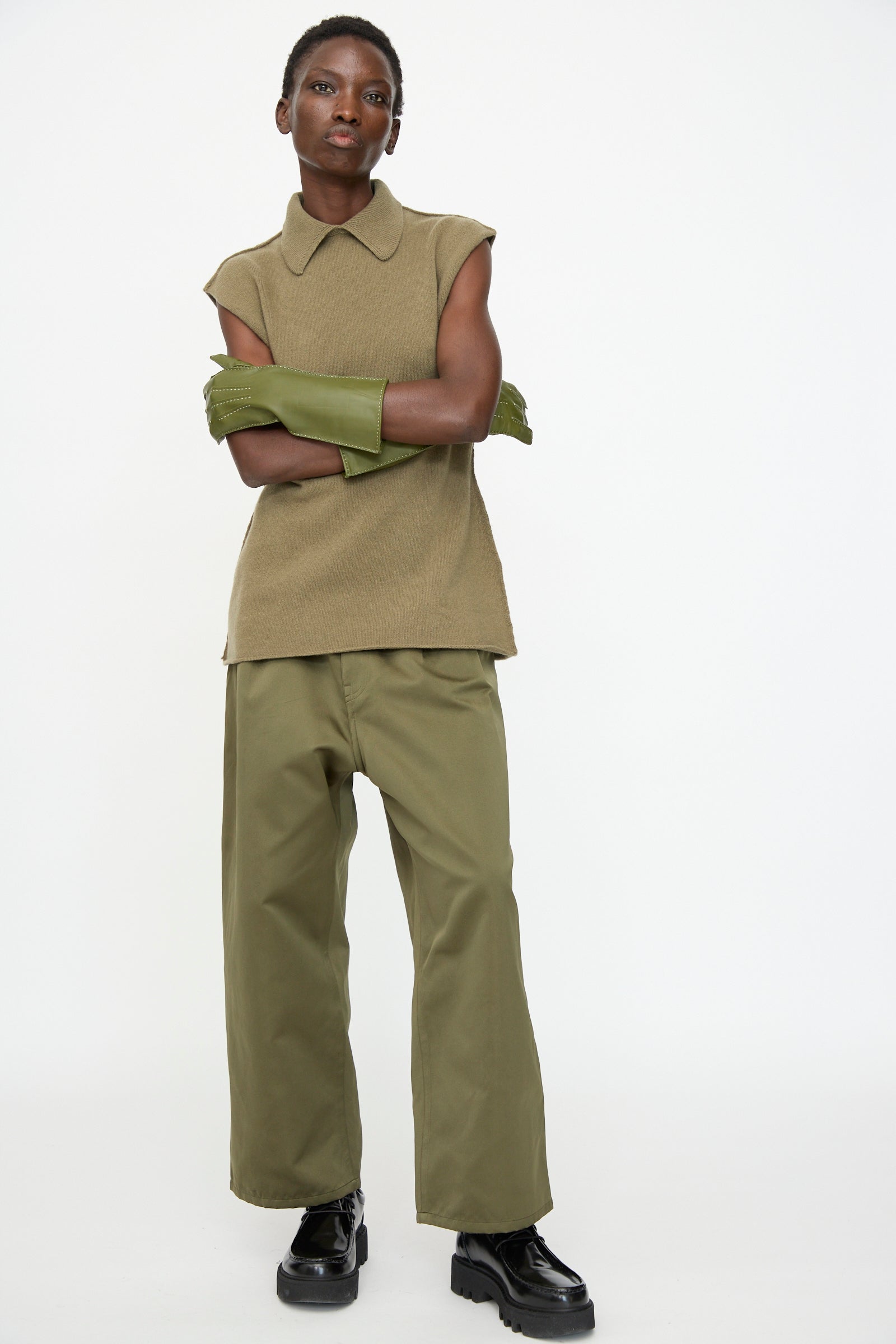 A person stands against a plain background wearing an olive green sleeveless top, wide-leg pants, Sofie D'Hoore's Leather Vasarely Gloves in Moss with silk lining, arms crossed, and styled with black platform shoes.