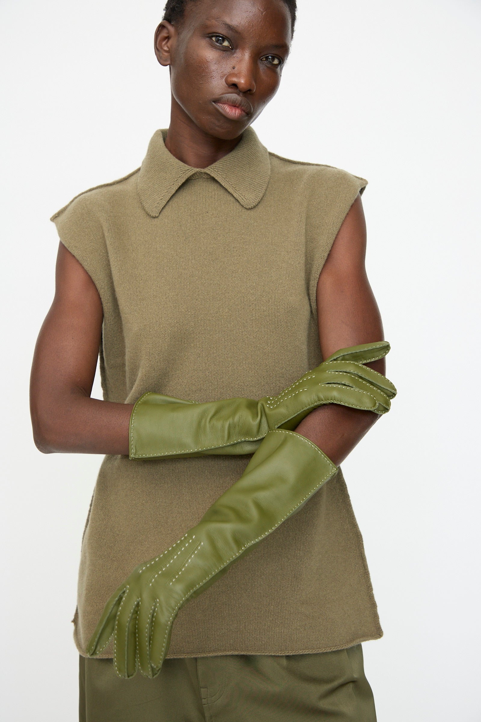 A person wearing an olive sleeveless top, olive pants, and Sofie D'Hoore Leather Vasarely Gloves in Moss stands against a plain light background with arms crossed.