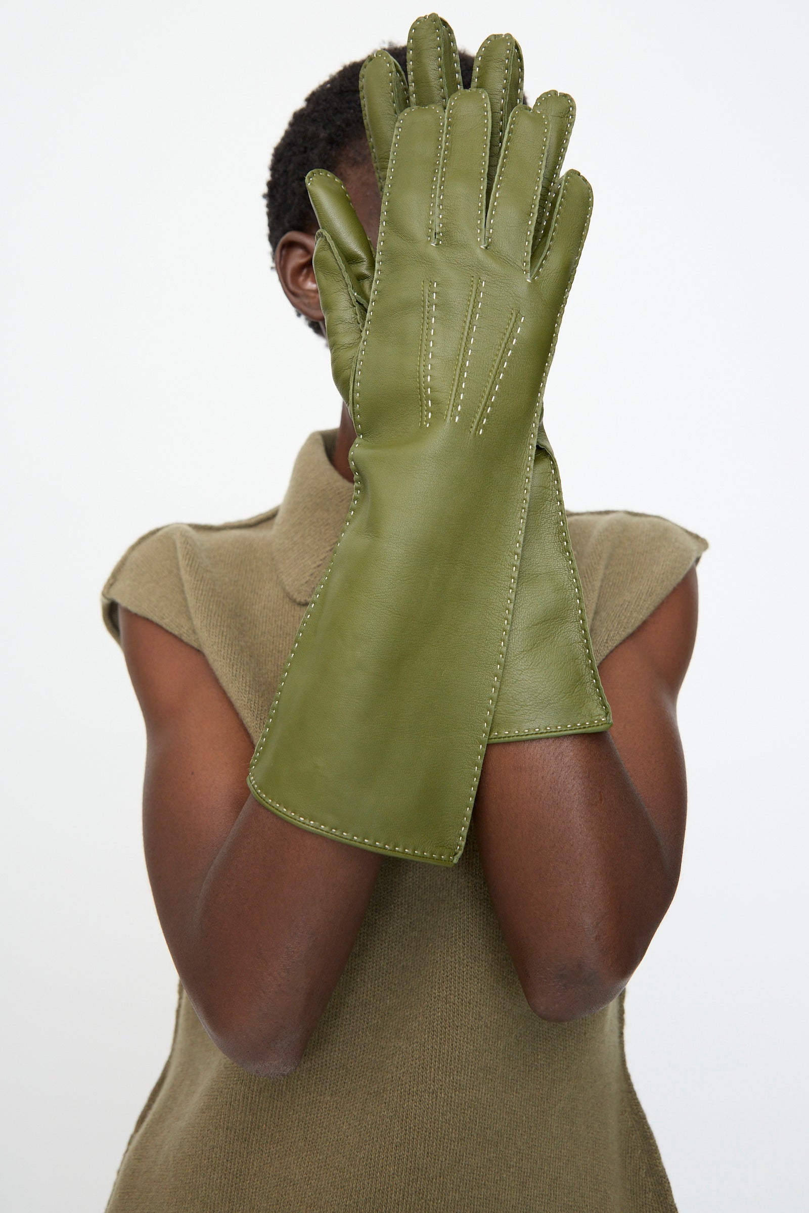 A person in a sleeveless olive top holds up Sofie D'Hoore's Leather Vasarely Gloves in Moss, partially covering their face against a plain background.