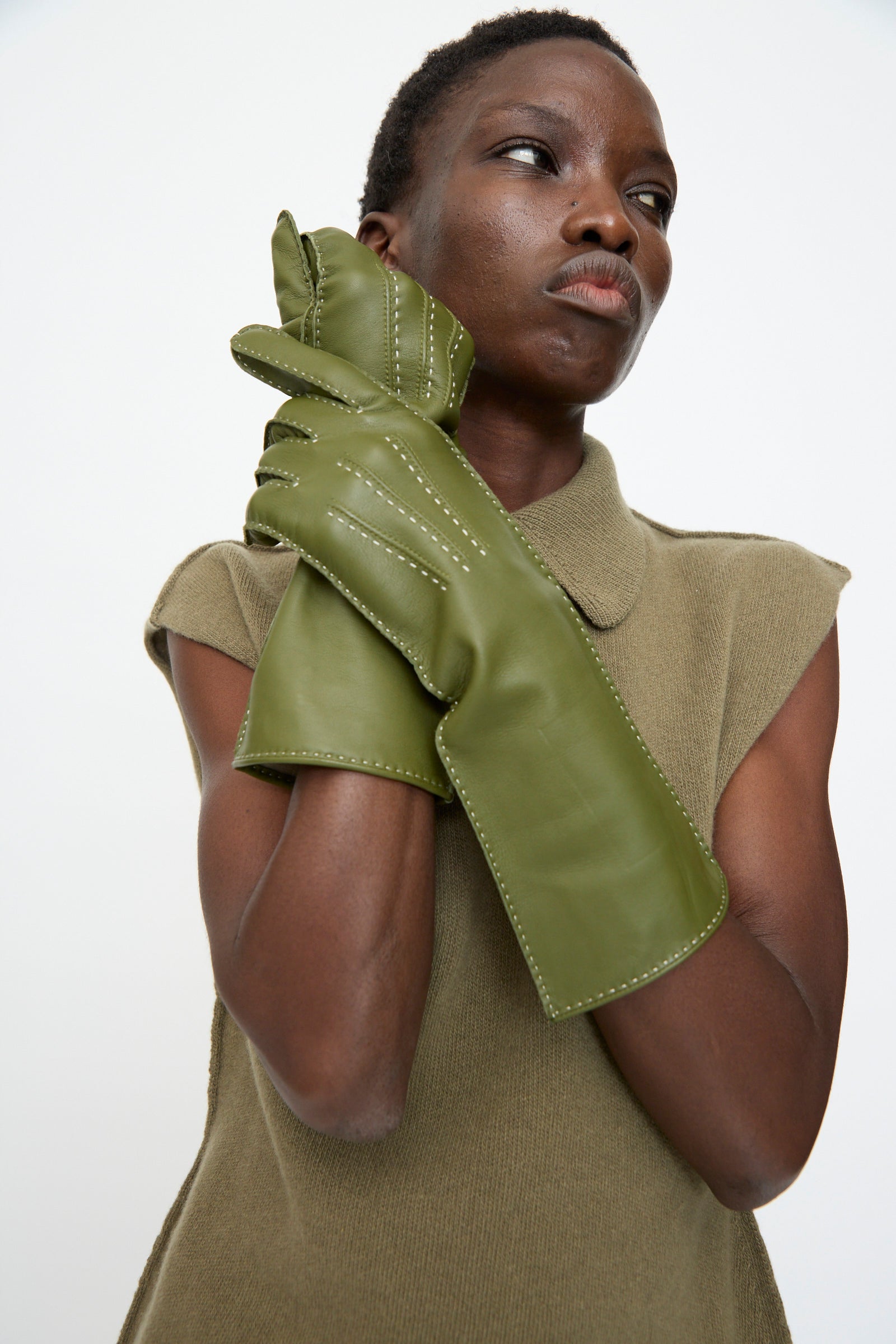A person wears an olive green sleeveless top and Sofie D'Hoore Leather Vasarely Gloves in Moss, made in France with silk lining, holding their gloved hands to their face while looking to the side.