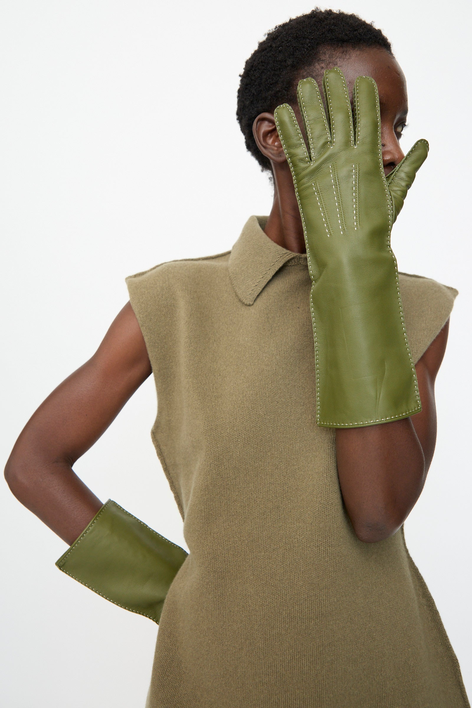 A person wears a sleeveless olive top and Sofie D'Hoore's Leather Vasarely Gloves in Moss—long French lamb leather gloves with silk lining—with one glove raised to partially cover their face.