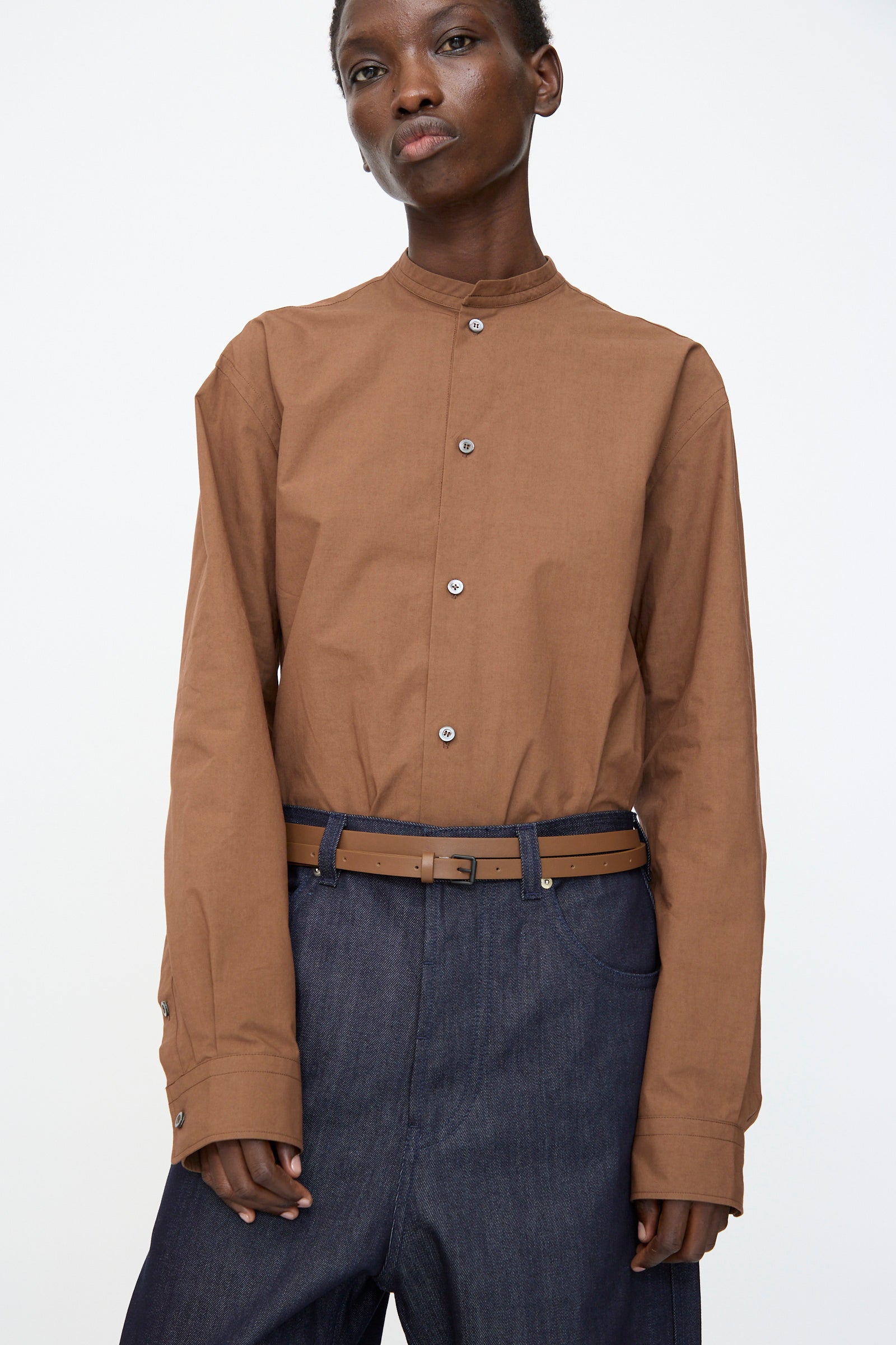 A person wears a brown mandarin-collar shirt, dark wide-leg jeans, and the Sofie D'Hoore Leather Vinck Belt in Chestnut with a matte black buckle, standing against a plain light background.