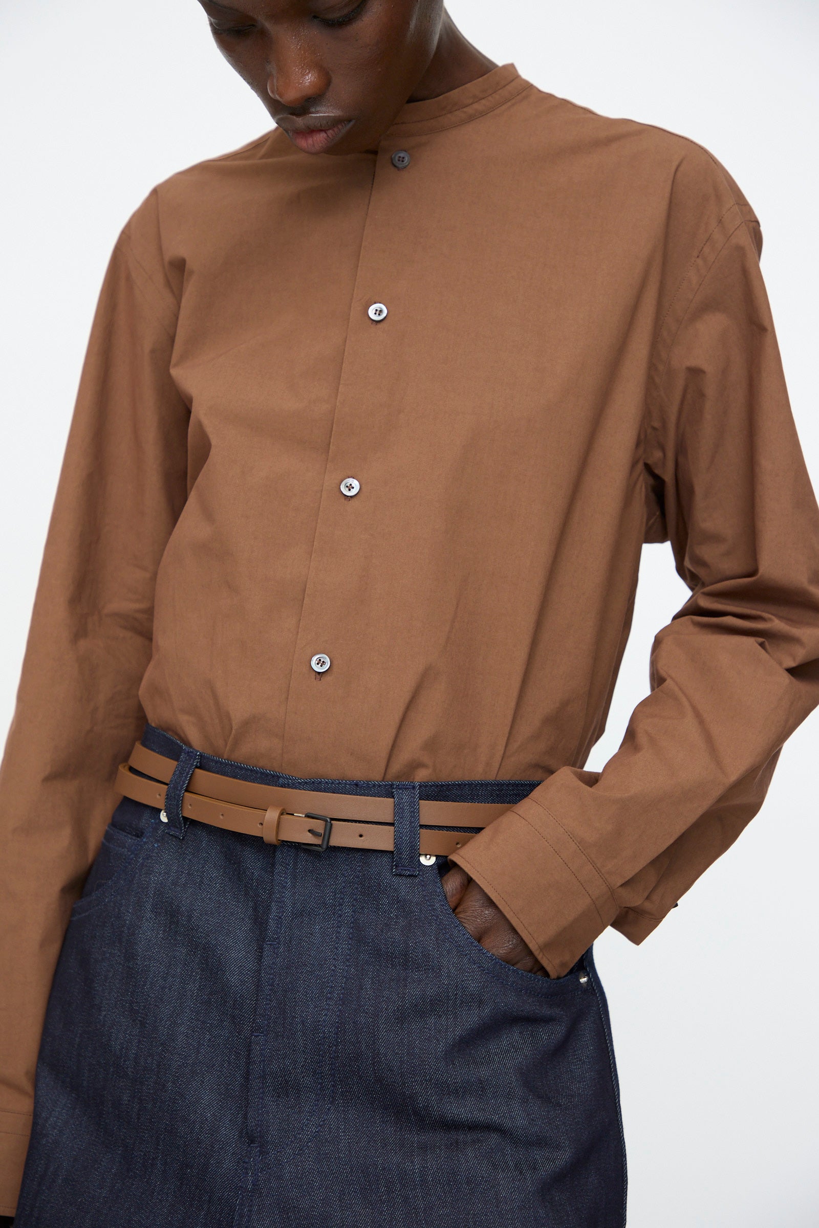 Model is shown from shoulders to hips wearing a brown button-up mandarin collar shirt, blue jeans, and the Sofie D'Hoore Leather Vinck Belt in Chestnut with a matte black buckle, one hand casually tucked in a pocket.