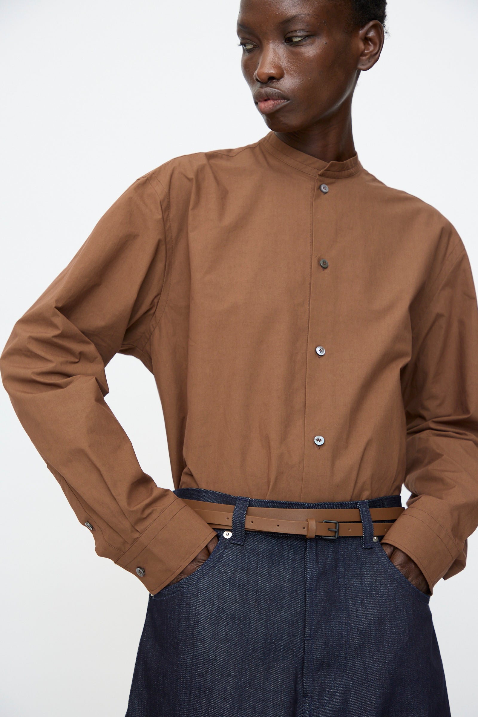 A person stands with hands in pockets, wearing a brown band-collar button-up shirt and dark blue high-waisted pants, accessorized with the Sofie D'Hoore Leather Vinck Belt in Chestnut featuring a matte black buckle against a plain background.
