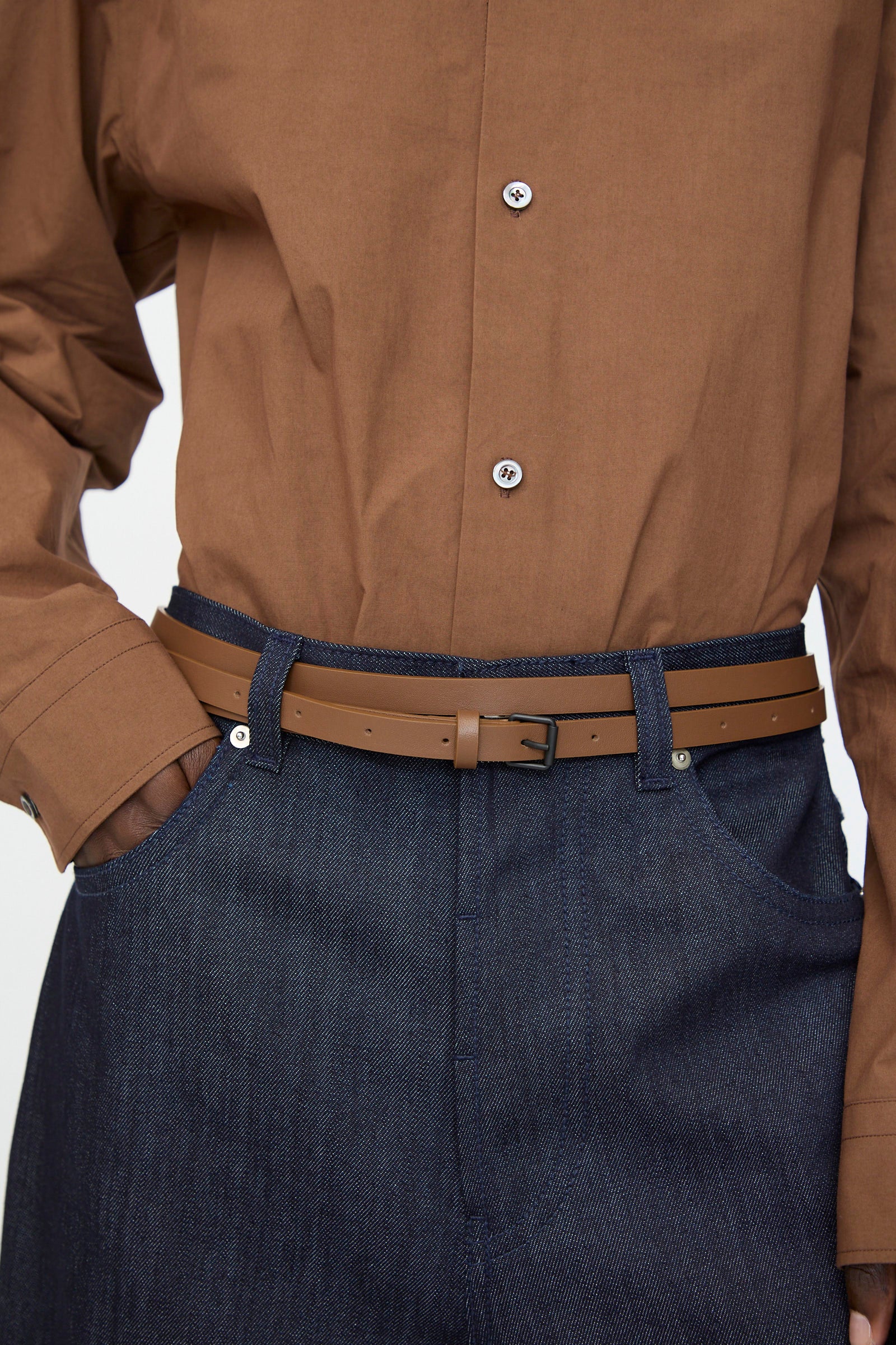 A person wearing a brown button-up shirt tucked into dark blue jeans with the Sofie D'Hoore Leather Vinck Belt in Chestnut featuring a matte black buckle.