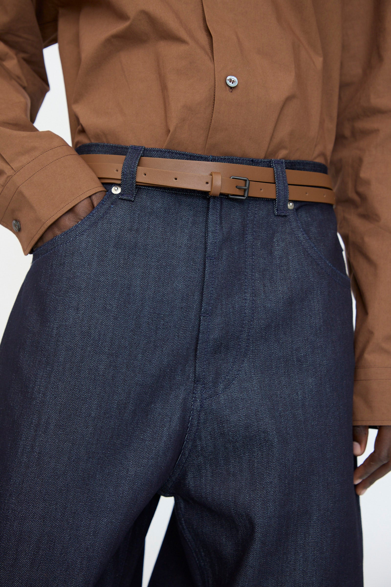 A person wearing a brown button-up shirt, dark denim jeans, and the Sofie D'Hoore Leather Vinck Belt in Chestnut with a matte black buckle, hands in their pockets.