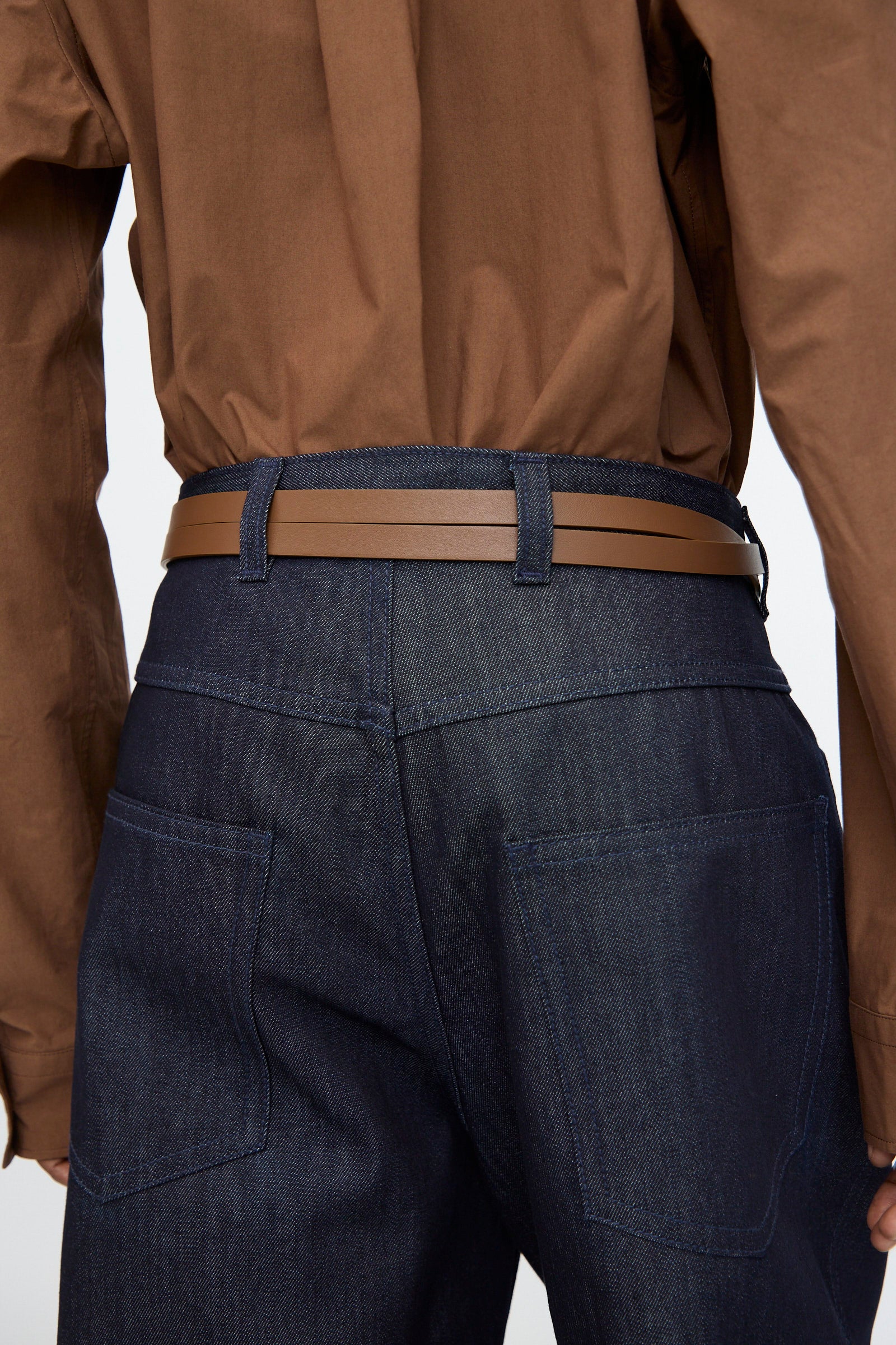 A person wears dark blue denim jeans and a tucked-in brown shirt, showcasing the Sofie D'Hoore Leather Vinck Belt in Chestnut with a matte black buckle; photographed from the back, highlighting the waistband and belt loops.