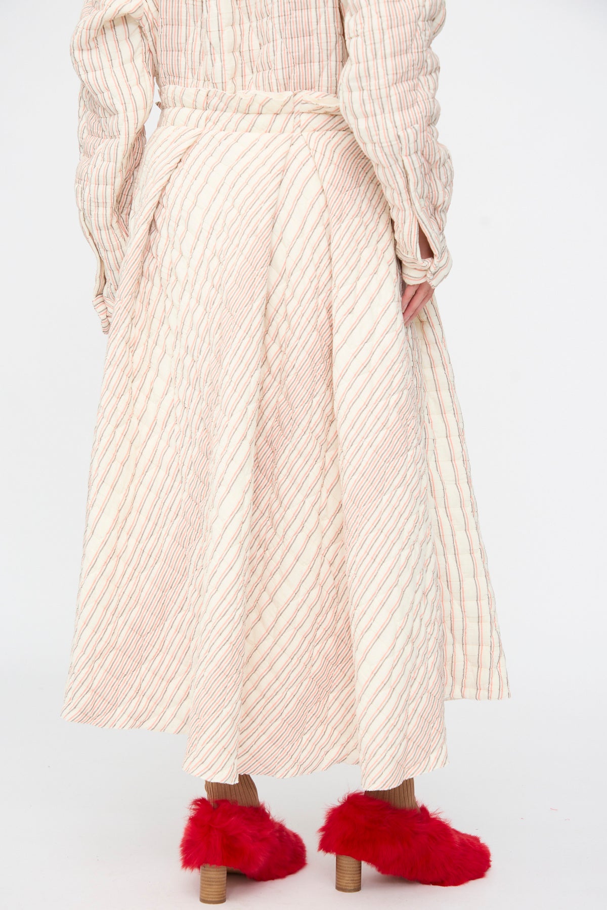 Rear view of a person wearing a cream-colored, quilted Sofie D’Hoore Quilted Cotton Savy Skirt in Red Stripe with thin red stripes and fuzzy red high-heeled shoes.