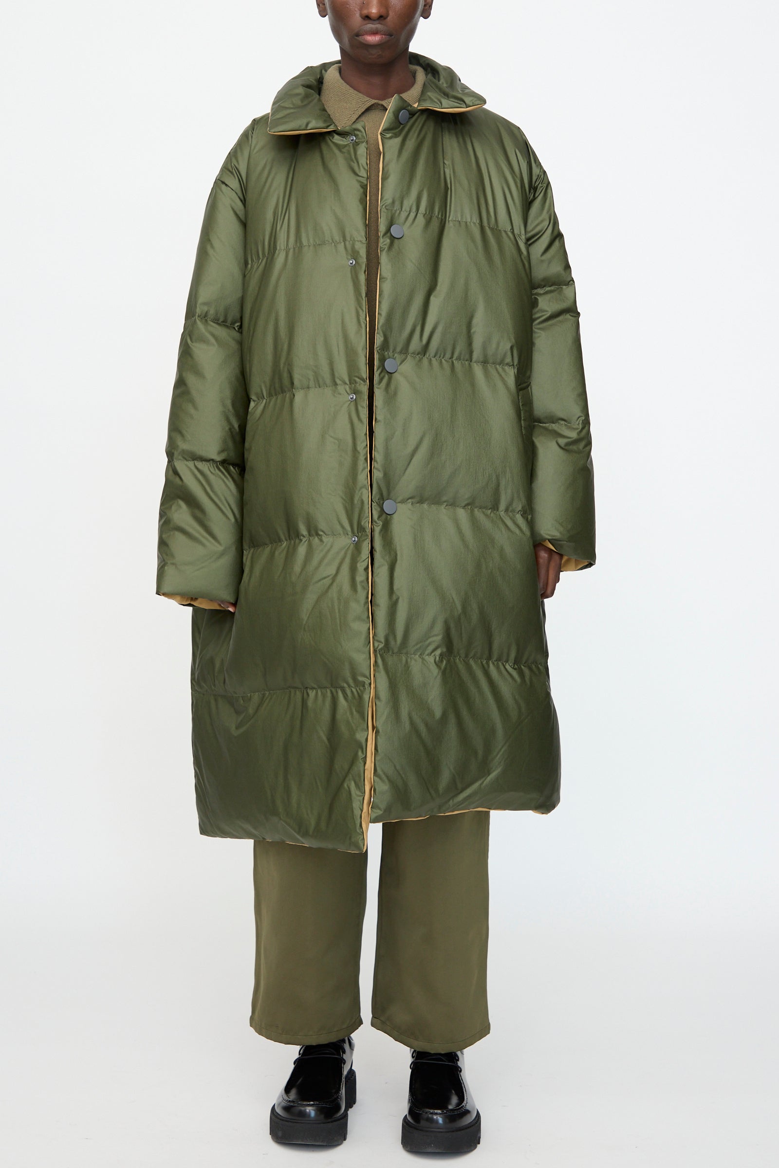 Model in Sofie D’Hoore’s Reversible Otis Down Coat in Mushroom and Khaki, styled with matching pants and black shoes, stands against a plain white background.