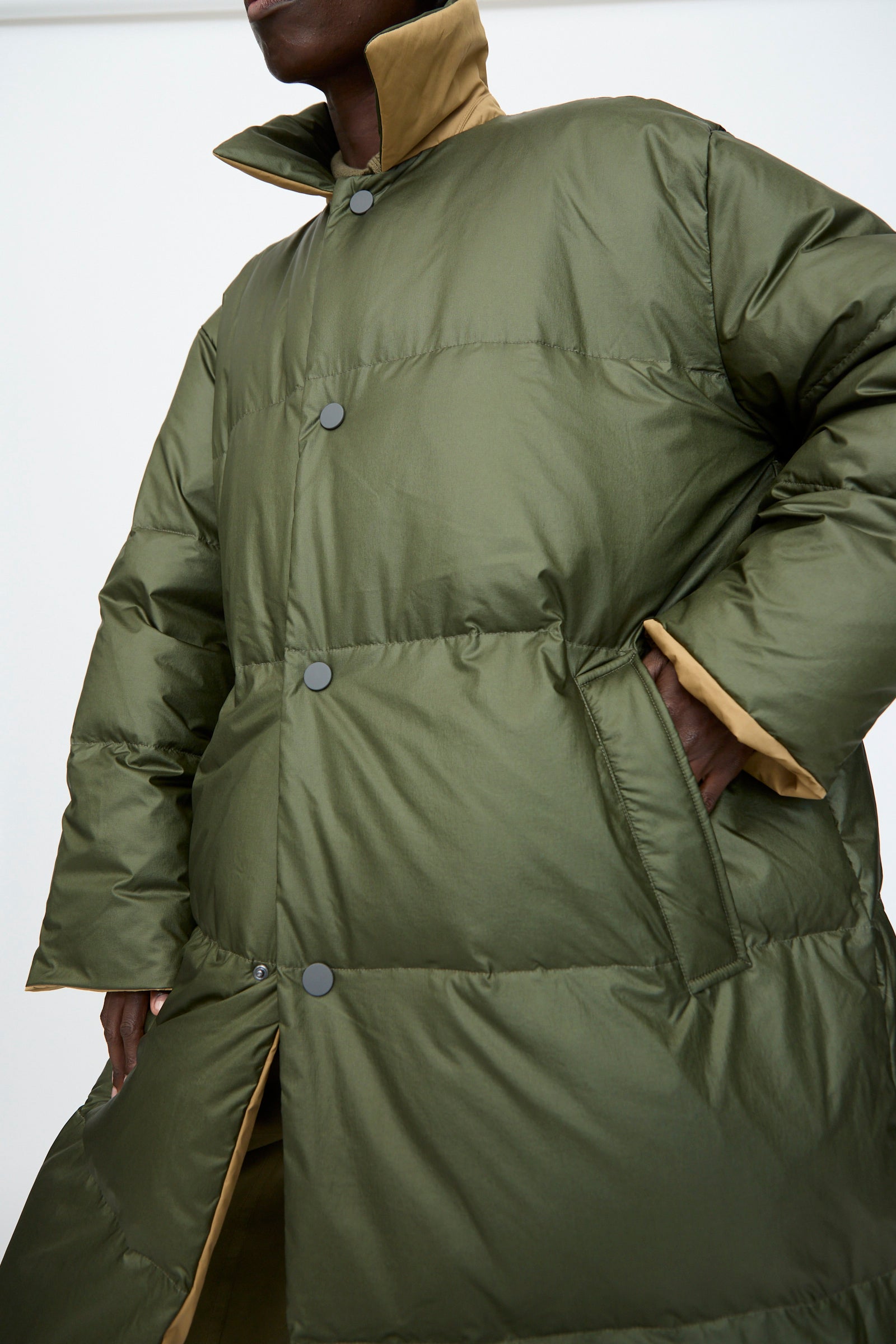 A person wearing the Sofie D'Hoore Reversible Otis Down Coat in Mushroom and Khaki, featuring a beige collar and snap buttons, has one hand in their pocket.