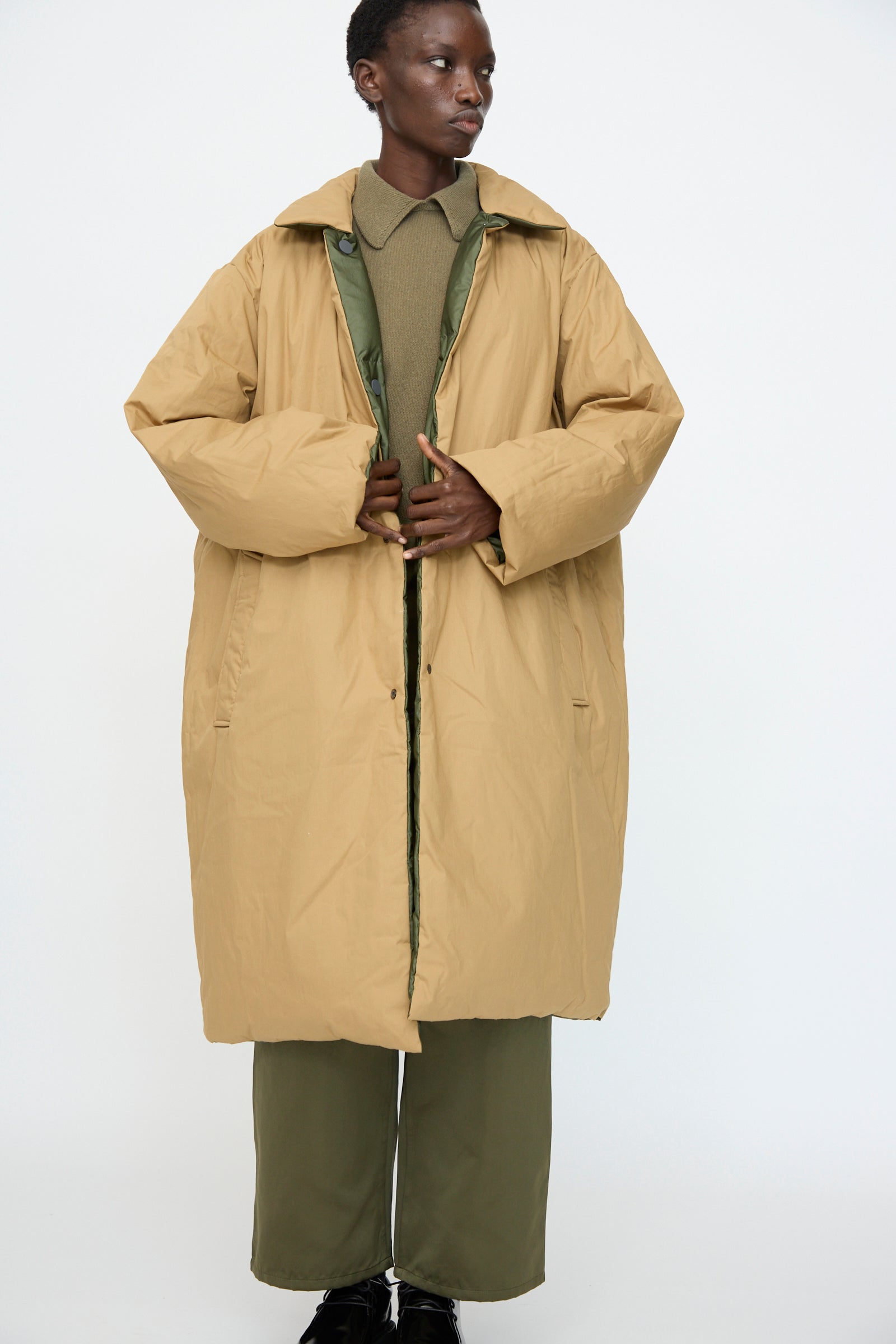 Model wears the Sofie D'Hoore Reversible Otis Down Coat in Mushroom and Khaki over a green outfit, standing against a plain white background.